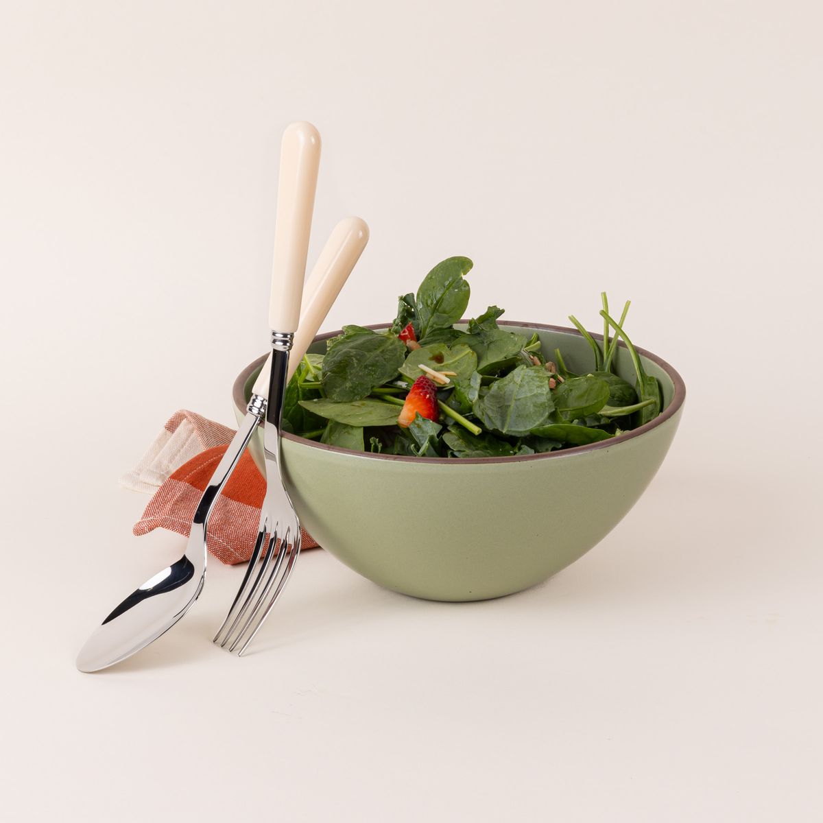 A serving size spoon and fork in a shiny steel with a matte cream handle leans against a sage green ceramic bowl filled with salad