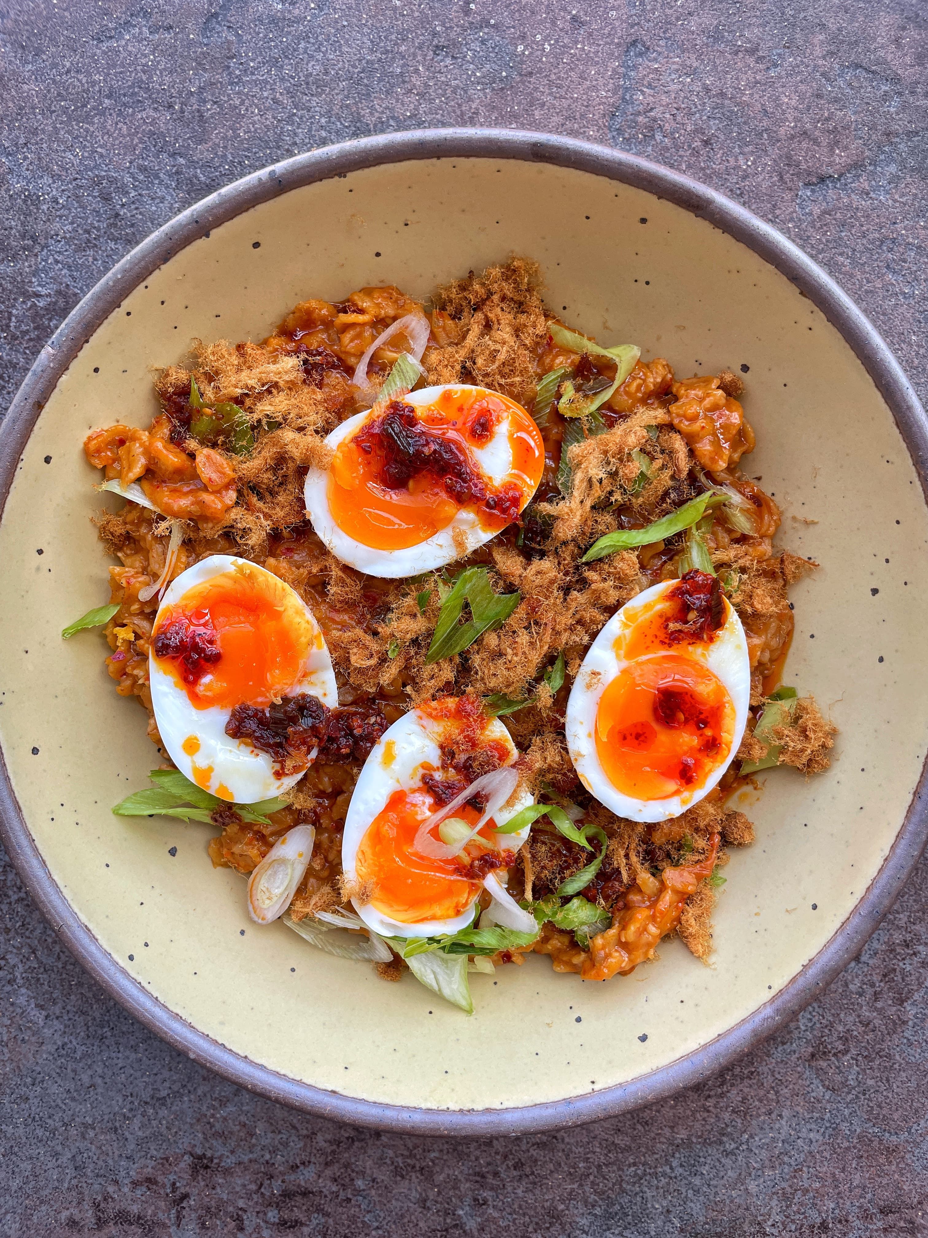 Closeup look of a meal in a ceramic bowl featuring oats, sliced eggs, chili crisp, and light veggies.