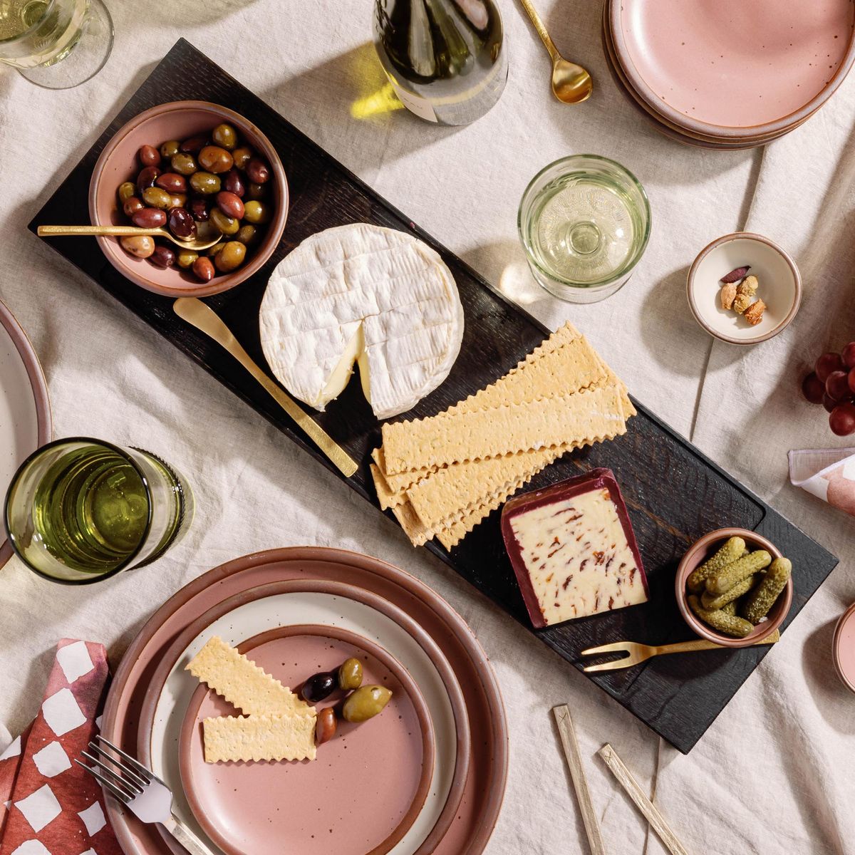 Ebonized wood cheese board with olives, crackers, brie, pickles, and wine glasses on a linen table, surrounded by pink plates and utensils.