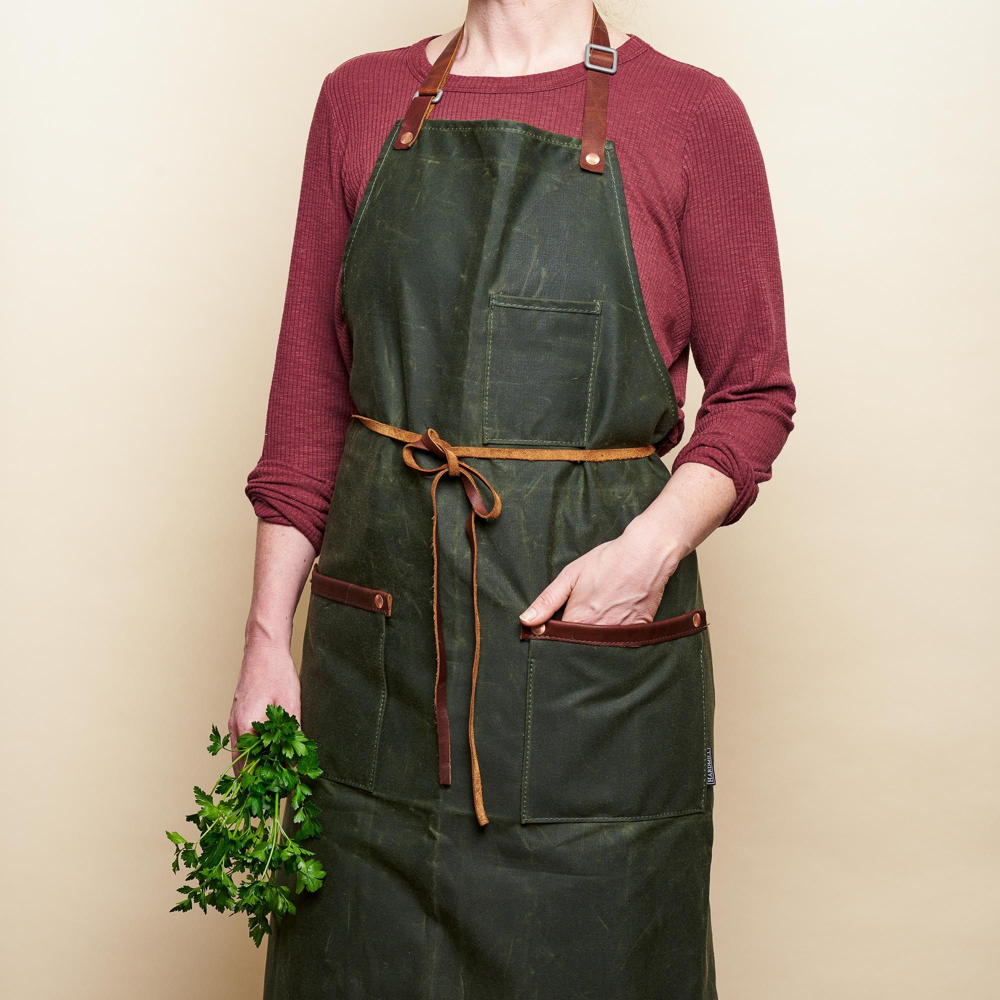 A woman models a forest-green waxed apron with leather straps and embellishments around the two wait pockets. There's a third pocket on the breast pocket. 