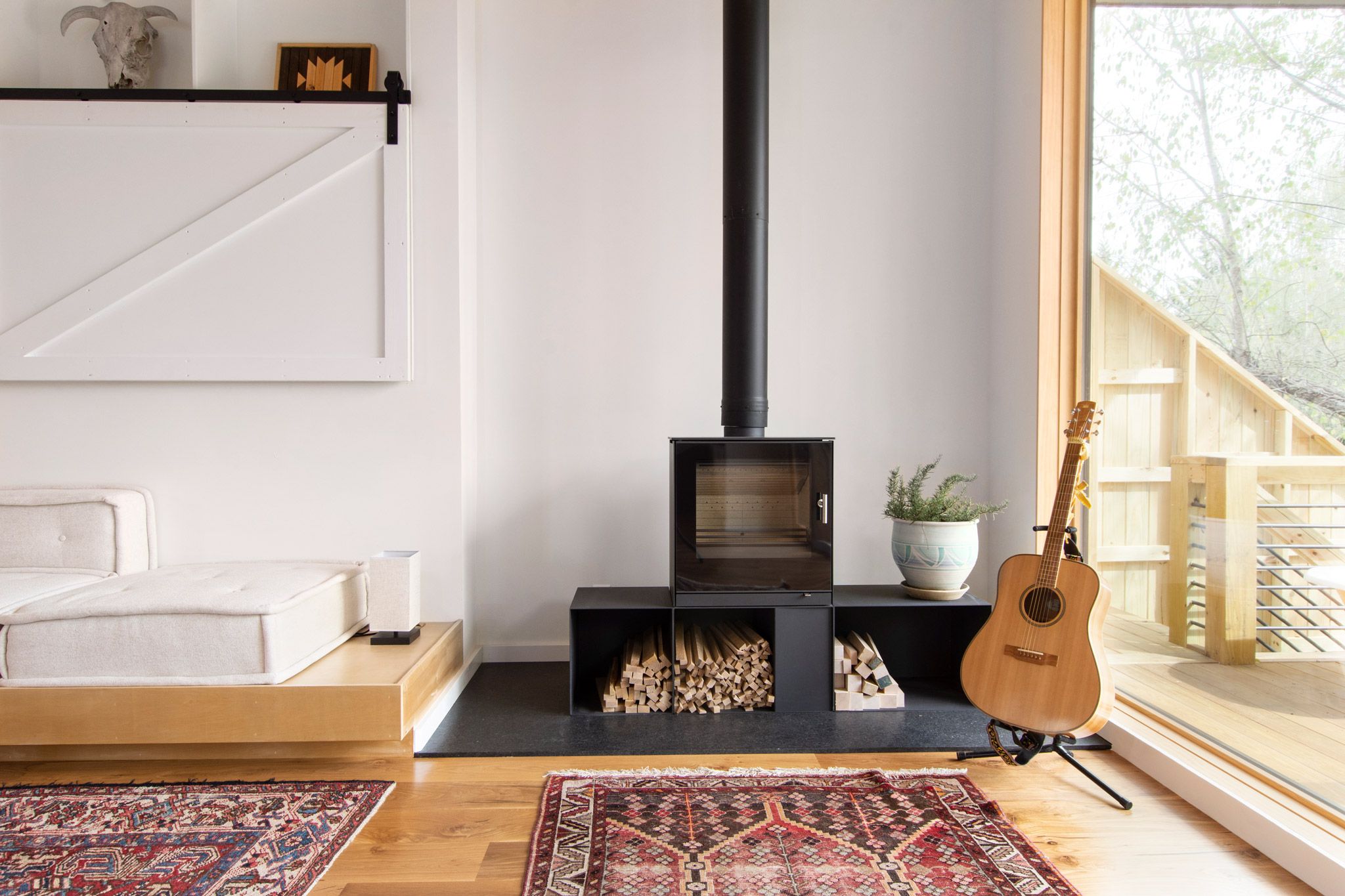 In a living room is a black minimal furnace with chopped wood below, and a guitar to the side.