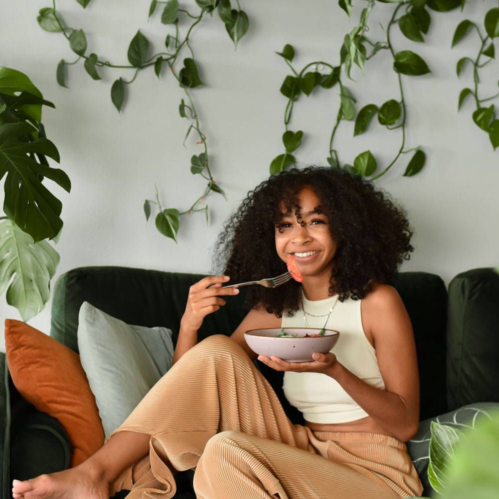 A person is smiling and eating from a shallow dinner bowl sitting on a couch with hanging greenery behind them.