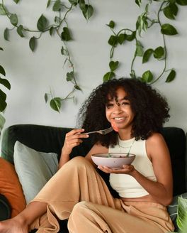 A person is smiling and eating from a shallow dinner bowl sitting on a couch with hanging greenery behind them.