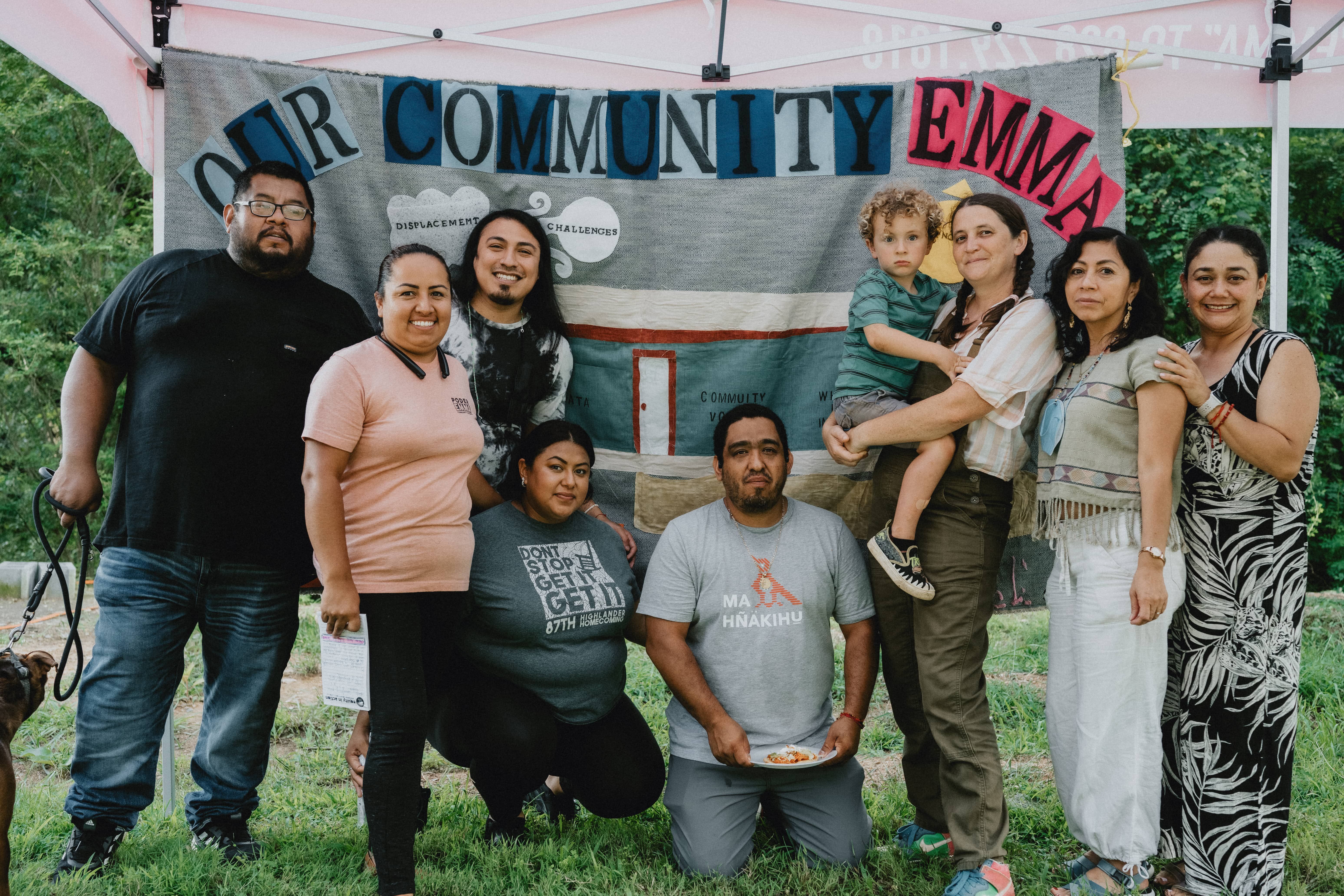 A group of people are gathered together smiling and outside standing in front of a tall banner that reads "Our Community Emma"