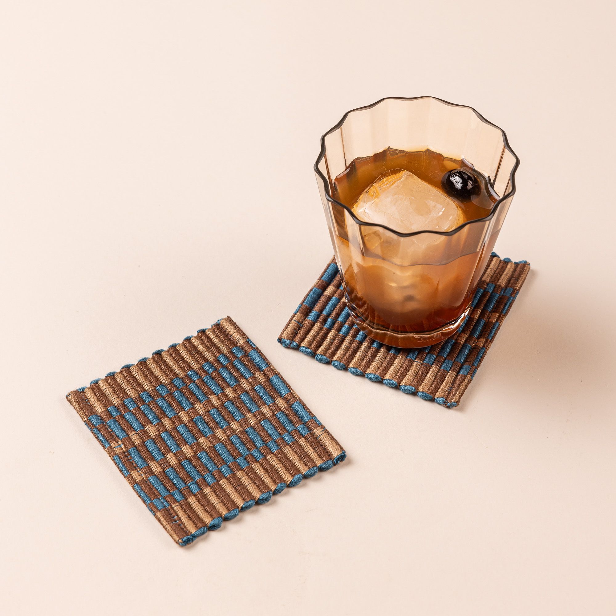 Two square coasters that are hand woven in a striped rectangular grid design in brown, tan, and blue colors. An elegant light tan lowball glass with a cocktail in it sits on the right coaster.