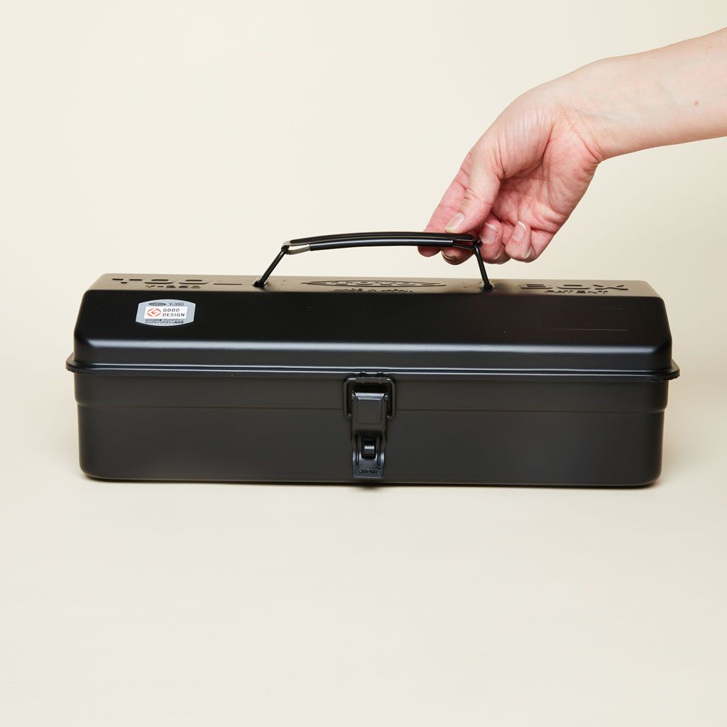 Fingers touch the black handle of a long black metal box with a lid and closure