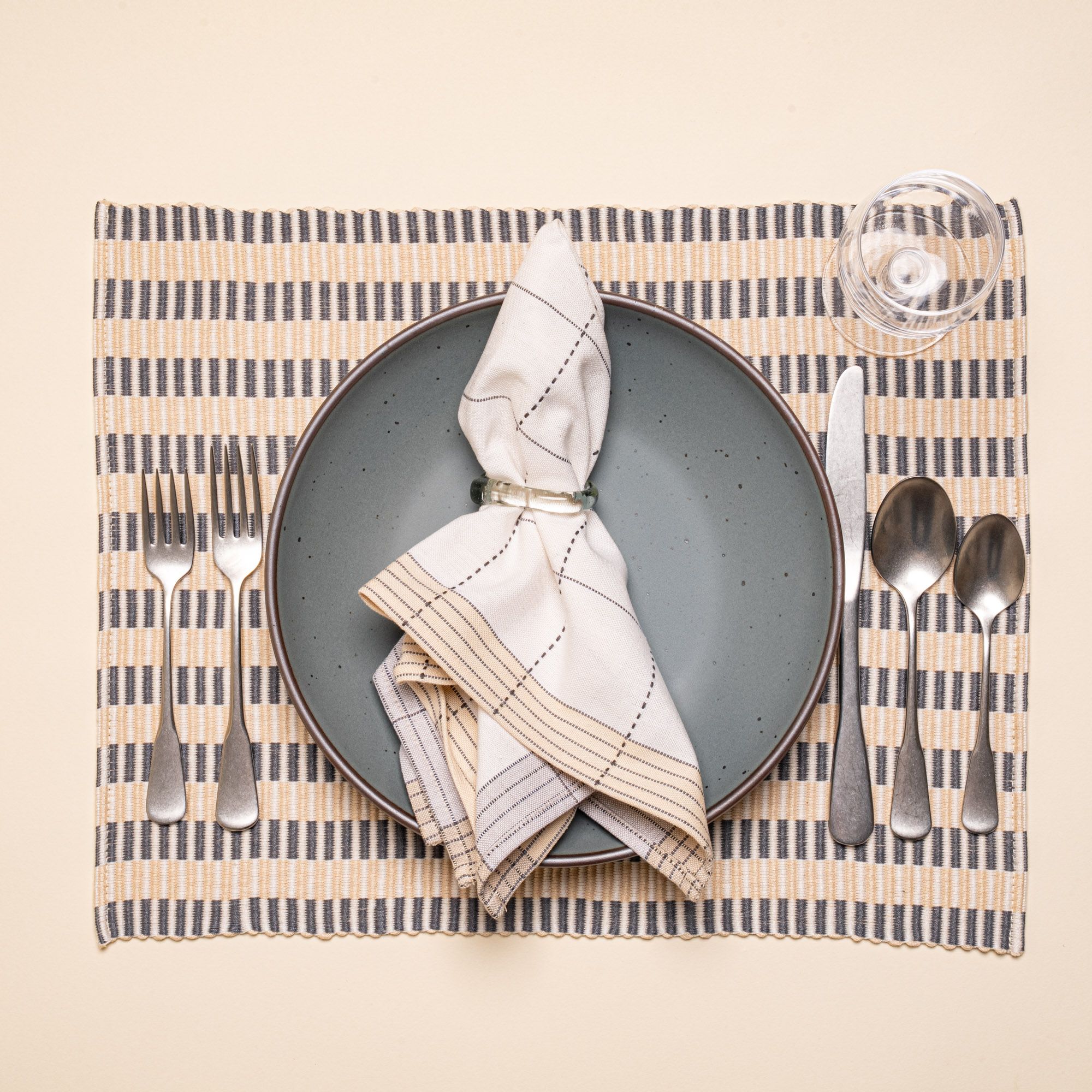 A white napkin with grey gridlines with light grey and natural striped edging on a blue-grey plate, with silverware and a glass
