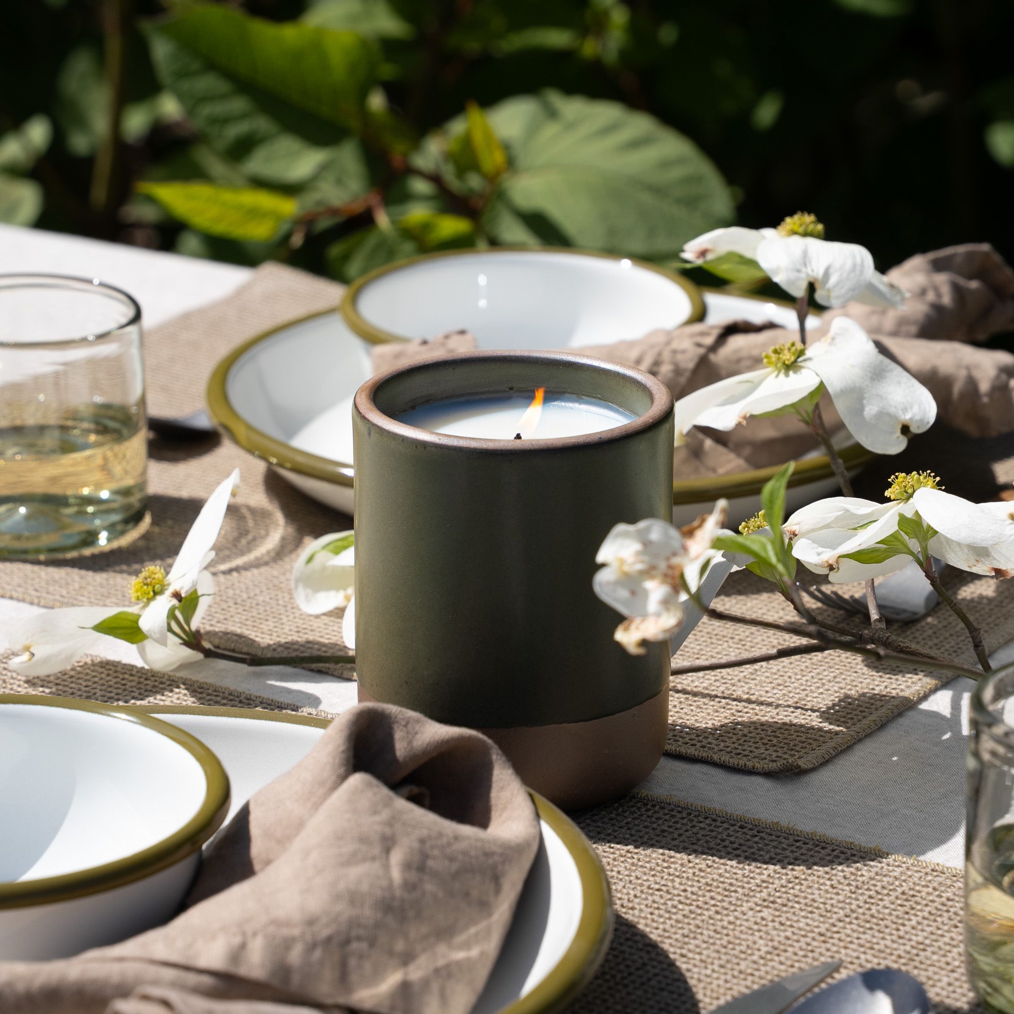 On an outdoor table, there are a few place settings and an outdoor candle in a ceramic cylindrical vessel in a dark forest green color that is lit.