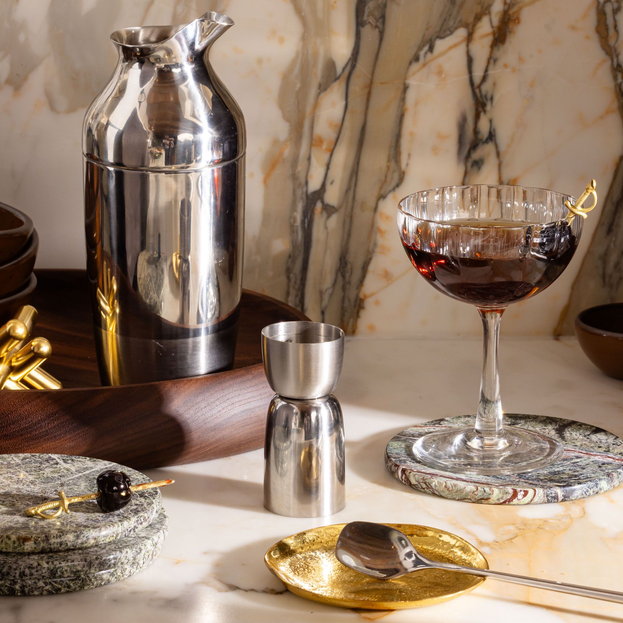 Cocktail setup with silver shaker, jigger, coupe glass with cherry garnish, and gold and stone coasters on marble.