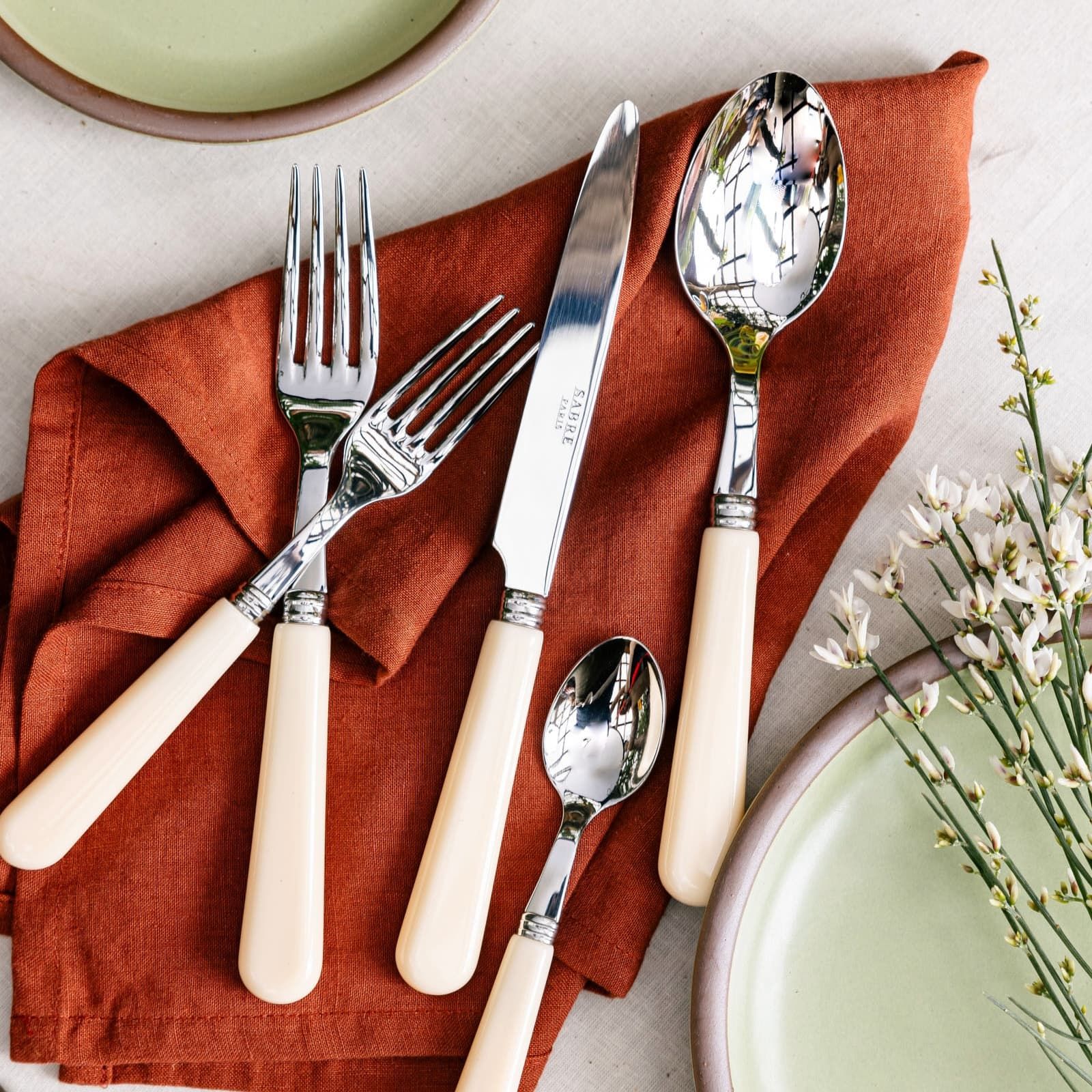 A five piece flatware set with shiny utensils and matte cream handles sit on a terracotta napkin surrounded by light greenery and sage green ceramic plates.