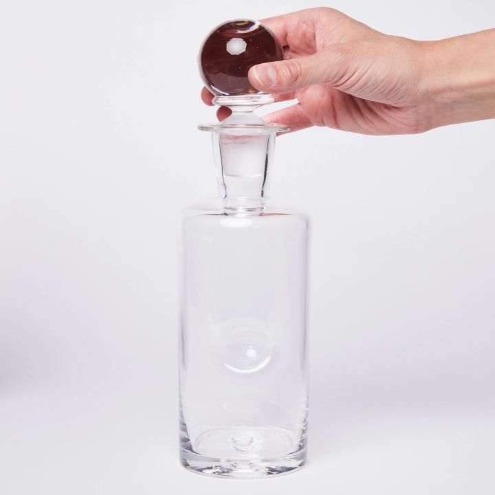 A hand holding a sphere-shaped stopper on top of a cylindrical clear glass decanter