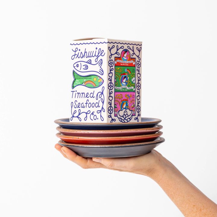 A hand holding up a stack of 4 ceramic plates in red, pink, blue and black colors, with an illustrated box of tinned fish on top.
