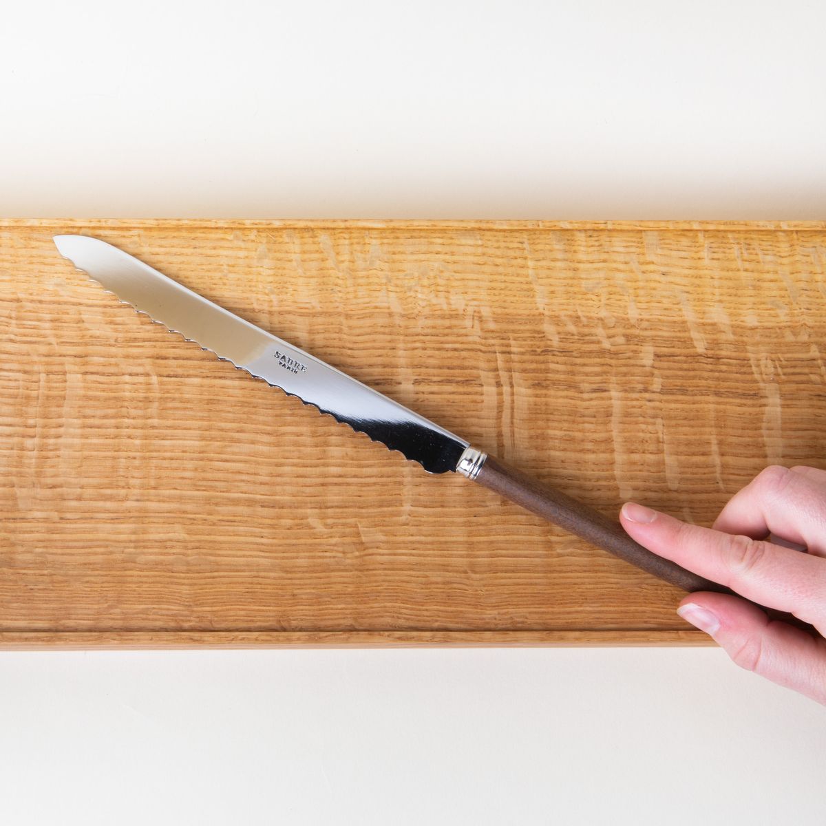 A hand holding a long bread knife with a steel blade and simple wood handle with a cutting board