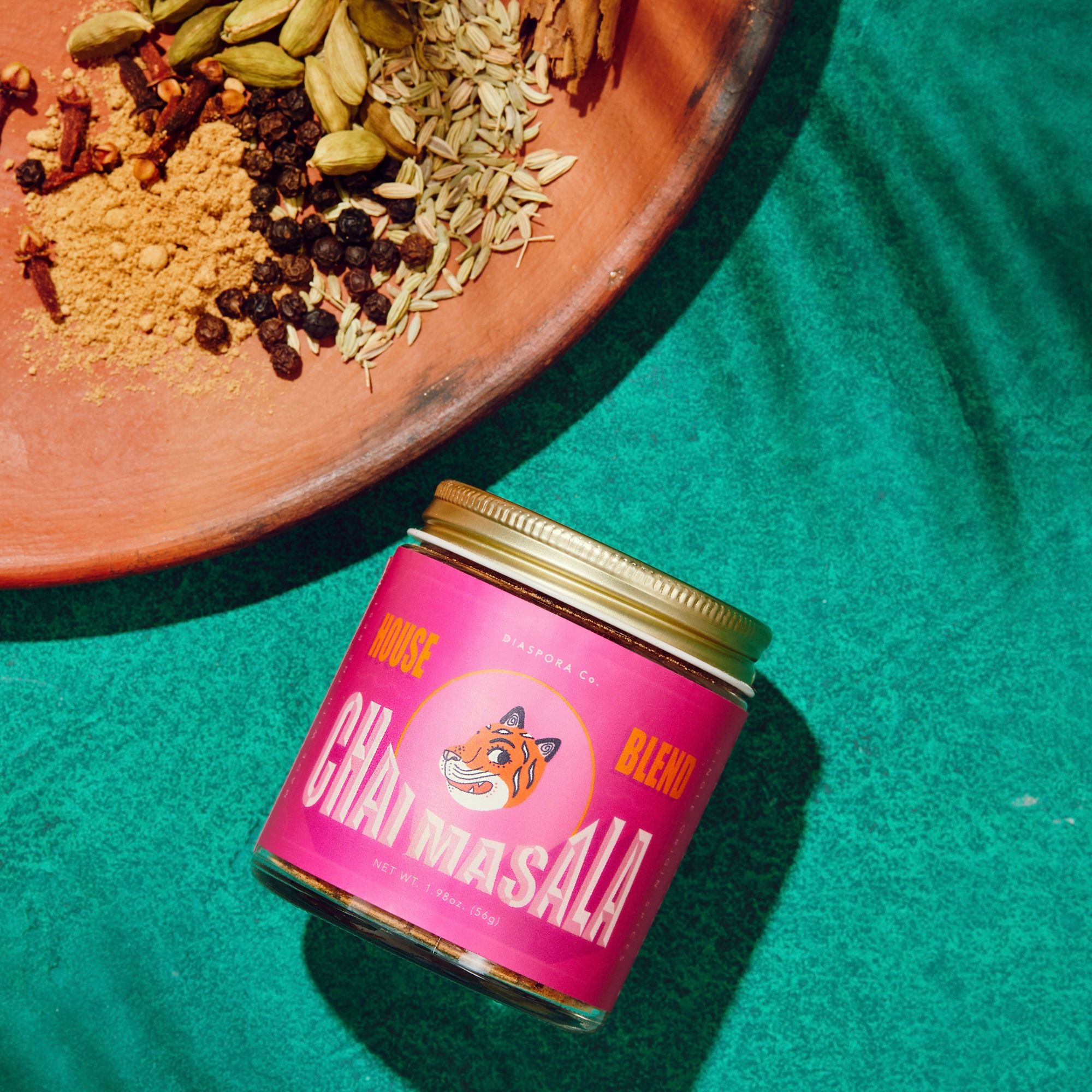 A jar wrapped with a hot pink label with a tiger illustration in the center with surrounding type that reads "House Blend Chai Masala", sits on a teal background next to a plate with spices