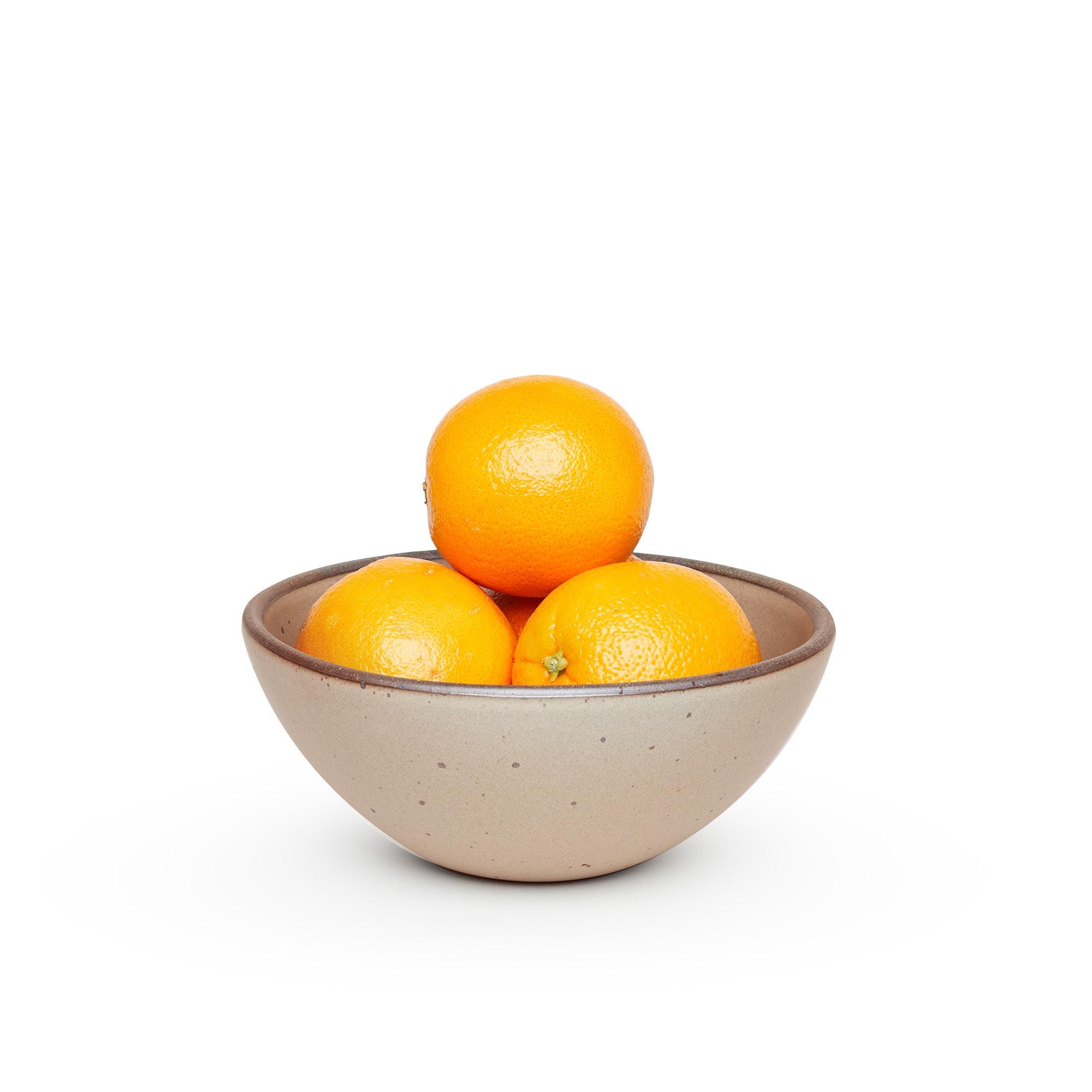A medium rounded ceramic bowl in a warm pale brown color featuring iron speckles and an unglazed rim, filled with oranges