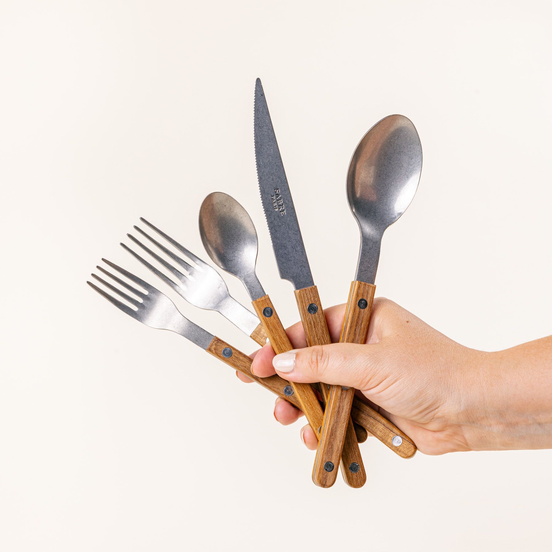 A hand holds a set of 5 flatware pieces with a teak wood handle: a salad fork, dinner fork, knife, spoon, teaspoon