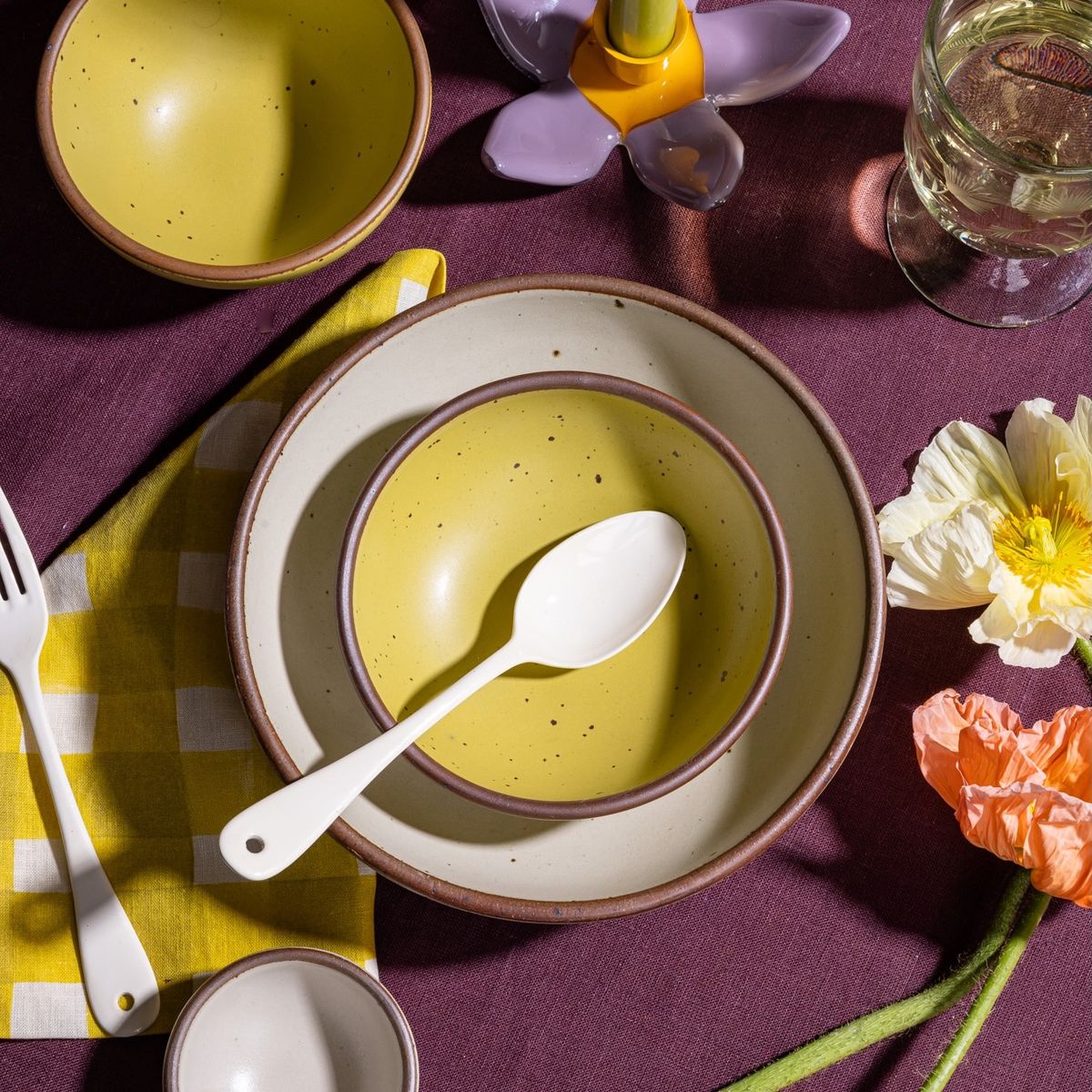 A dinner spoon sitting on a zesty yellow shallow bowl and cream plate, surrounded by flowers and printed yellow napkin.