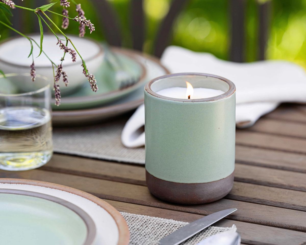 On an outdoor table, there is a few place settings and an outdoor candle in a ceramic cylindrical vessel in a sage green color that is lit.