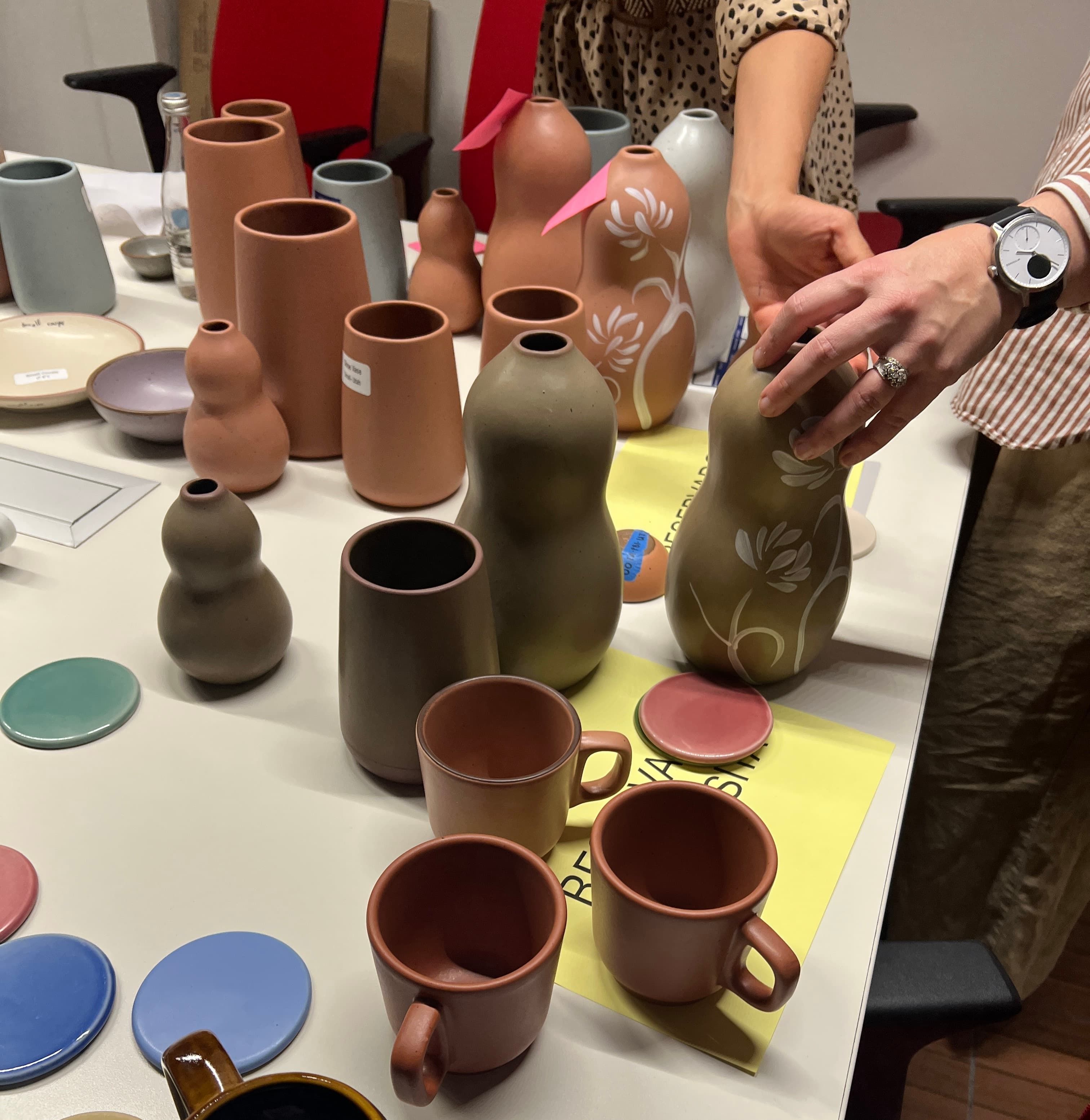 Two hands inspect vases in neutral clay and olive tones on a white tabletop surrounded by mug and color samples.