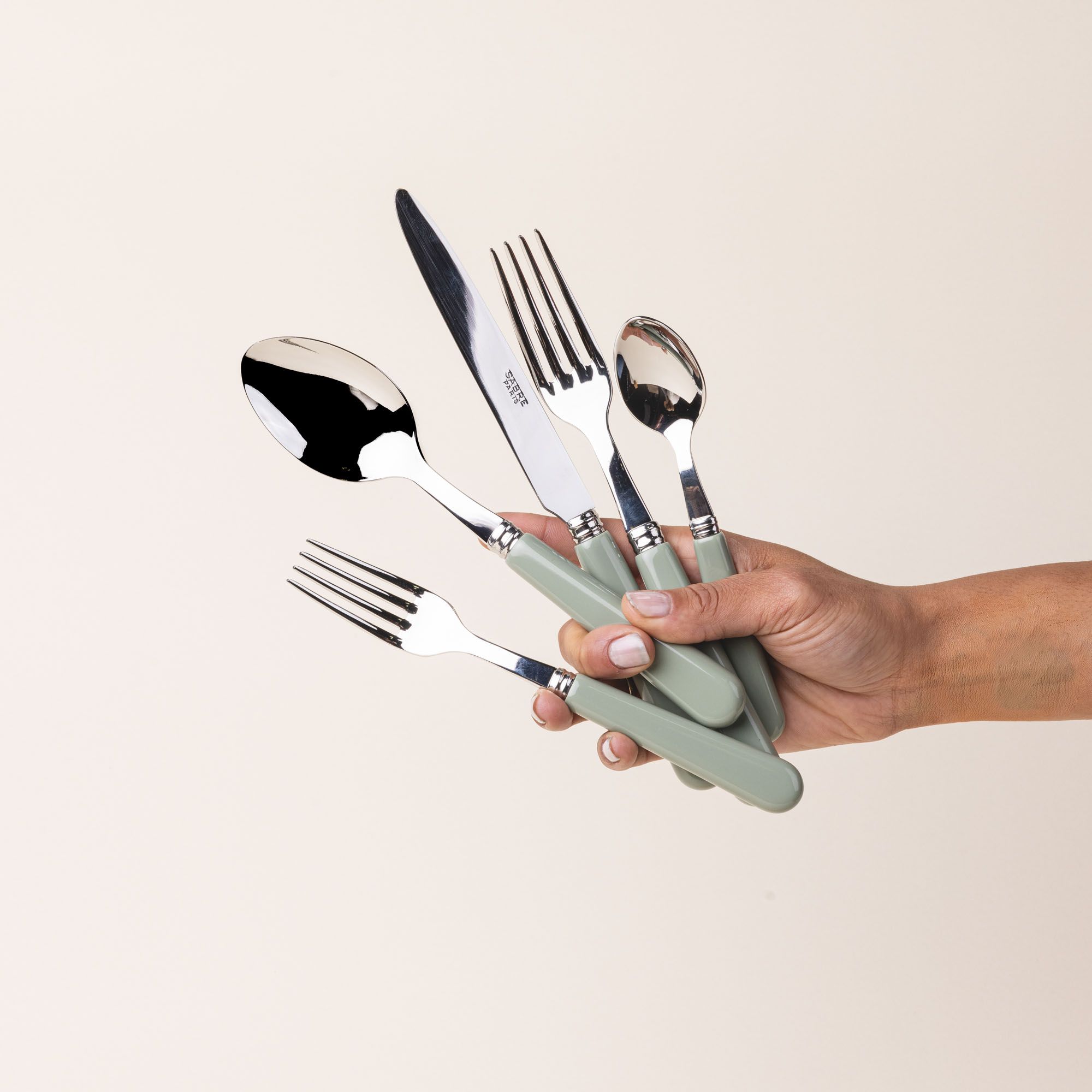 A hand holds a five piece flatware set with shiny utensils and matte sage green handles