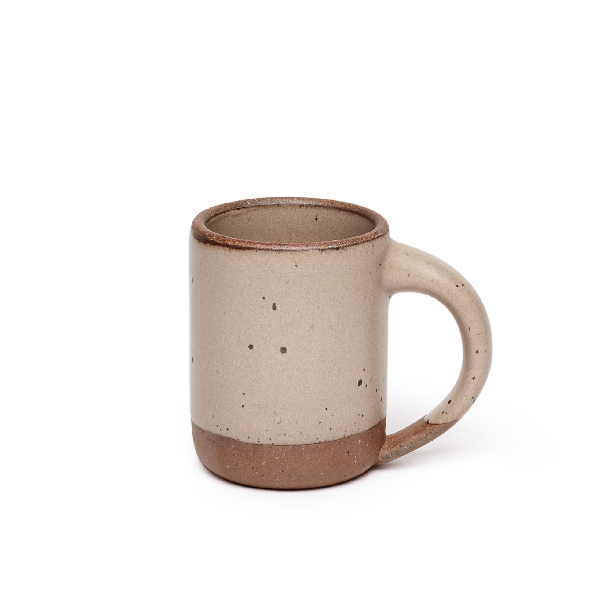 A medium sized ceramic mug with handle in a warm pale brown color featuring iron speckles and unglazed rim and bottom base.