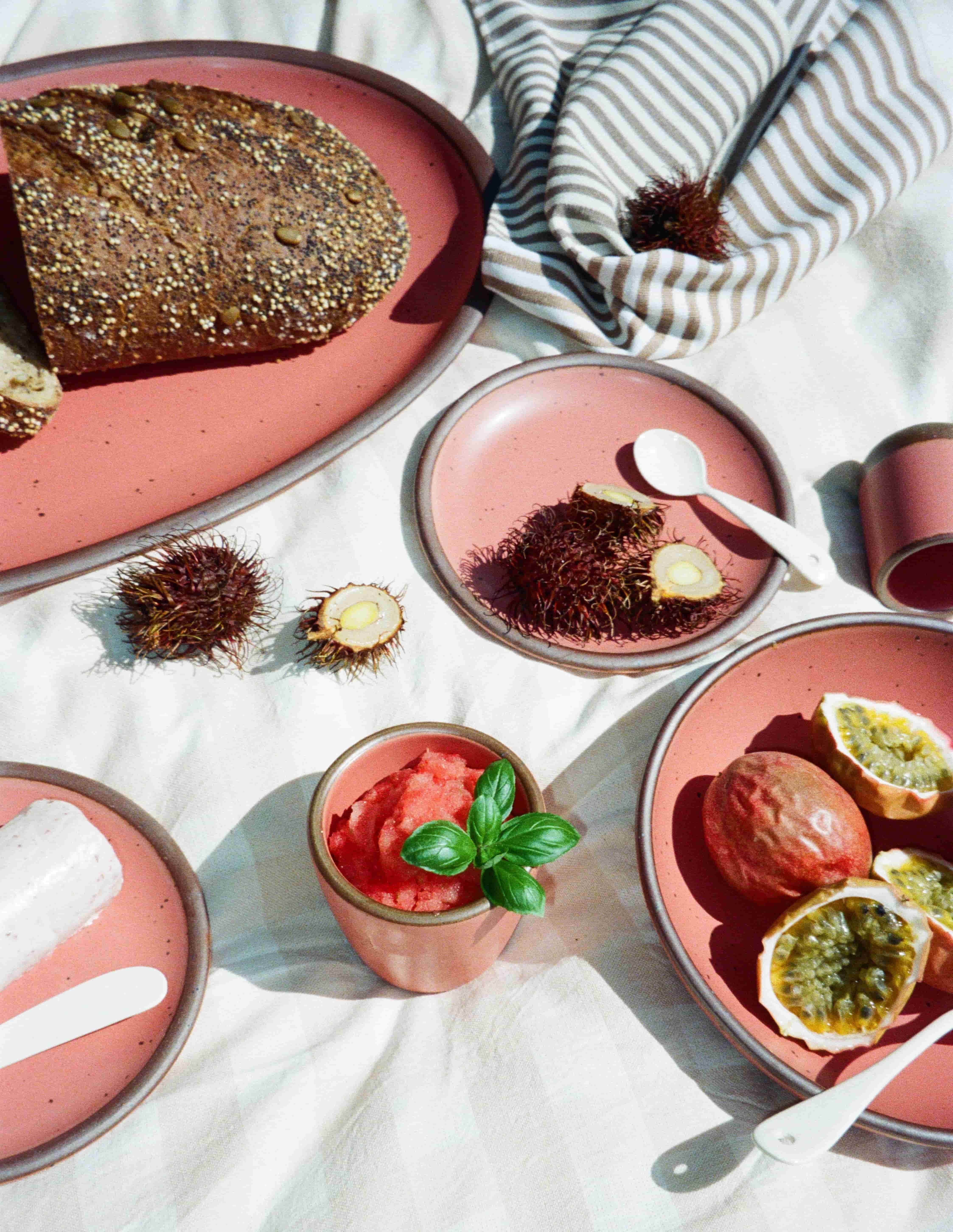 Sliced passion fruit, rambutan, watermelon granita, and seeded bread on pink plates atop a cream-striped picnic blanket.