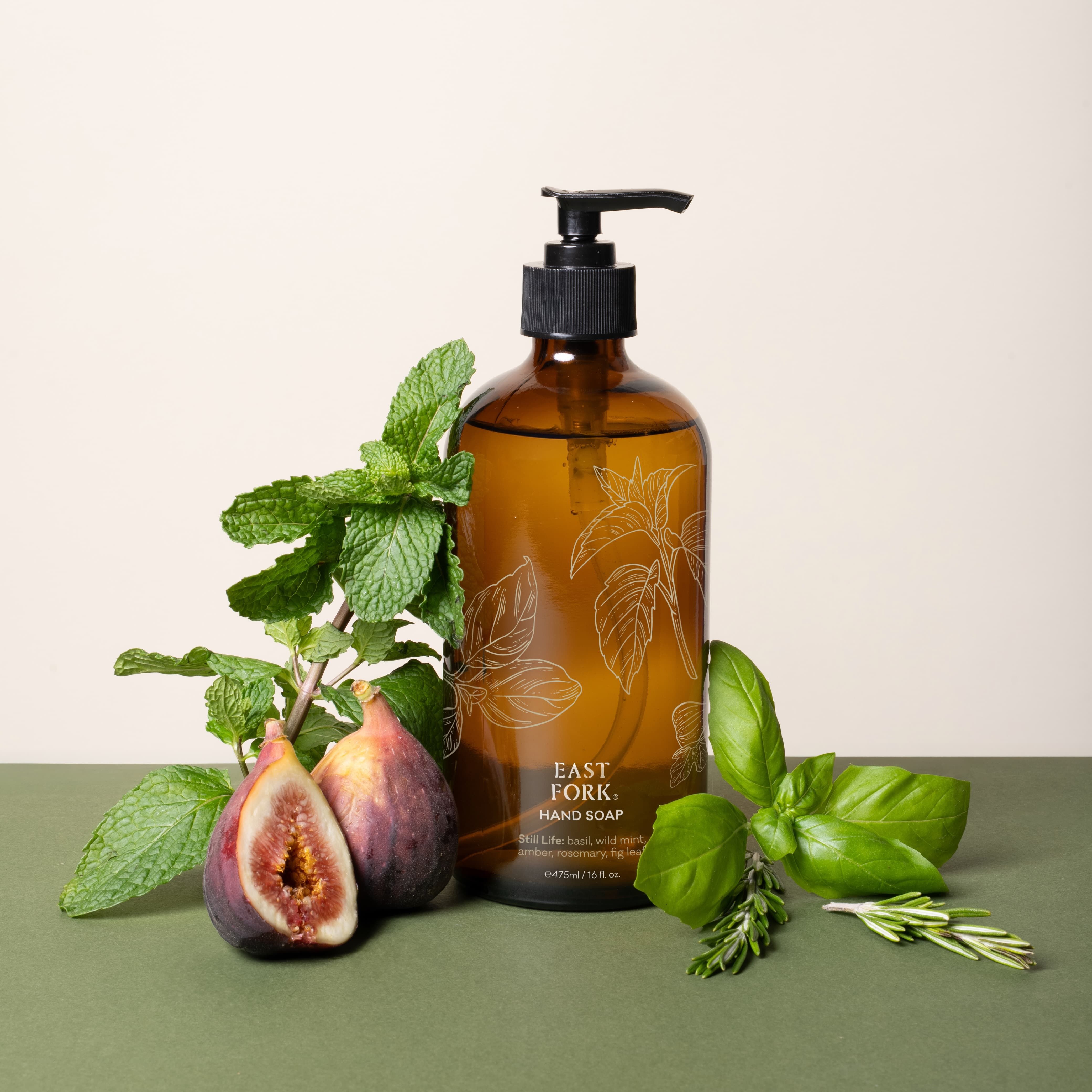 A brown glass bottle of East Fork Hand Soap labeled “Still Life” is surrounded by fresh basil, mint, rosemary, and figs on a green and cream backdrop.