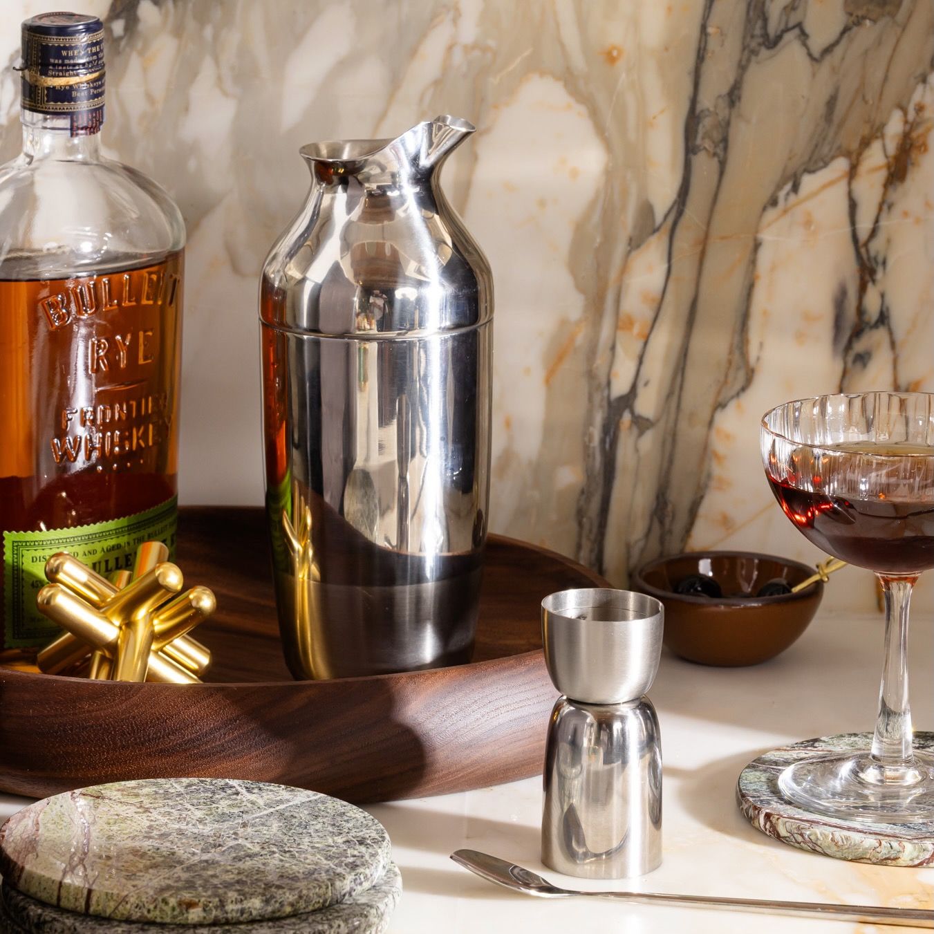 On a counter, a bar set up with a stainless steel cocktail shaker, measurer, whiskey, coasters and fluted coupe glass, against a marble backsplash.