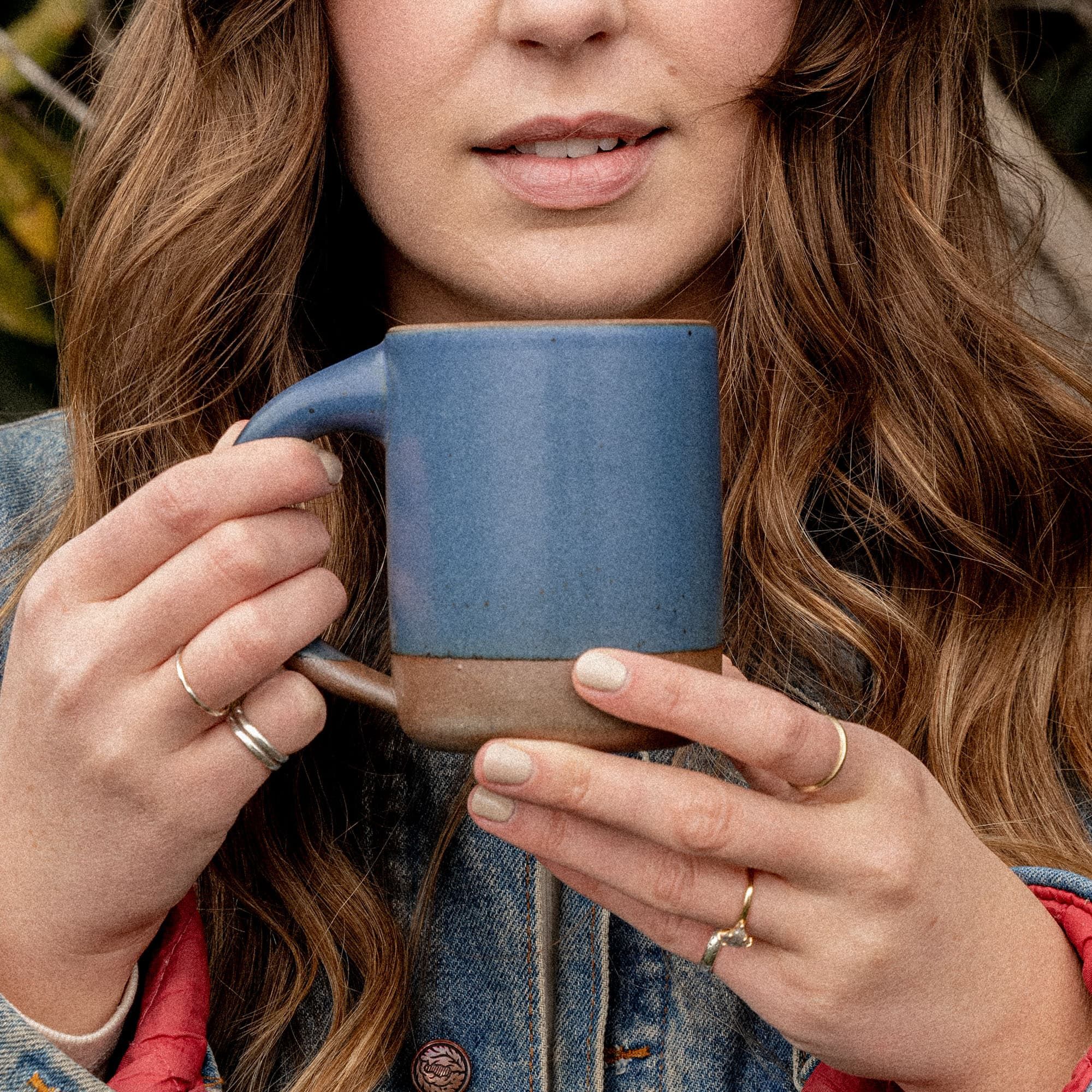 A woman holding The Mug in a cool medium blue color with two hands near her face outside.