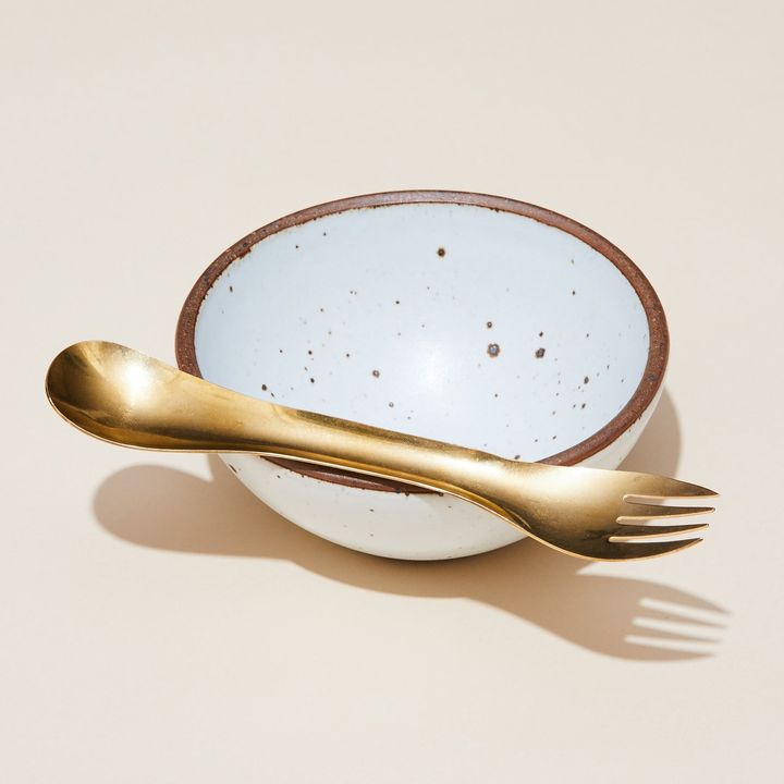 A brass eating utensil with spoon at one end and fork at the other, balanced atop an East Fork Breakfast Bowl