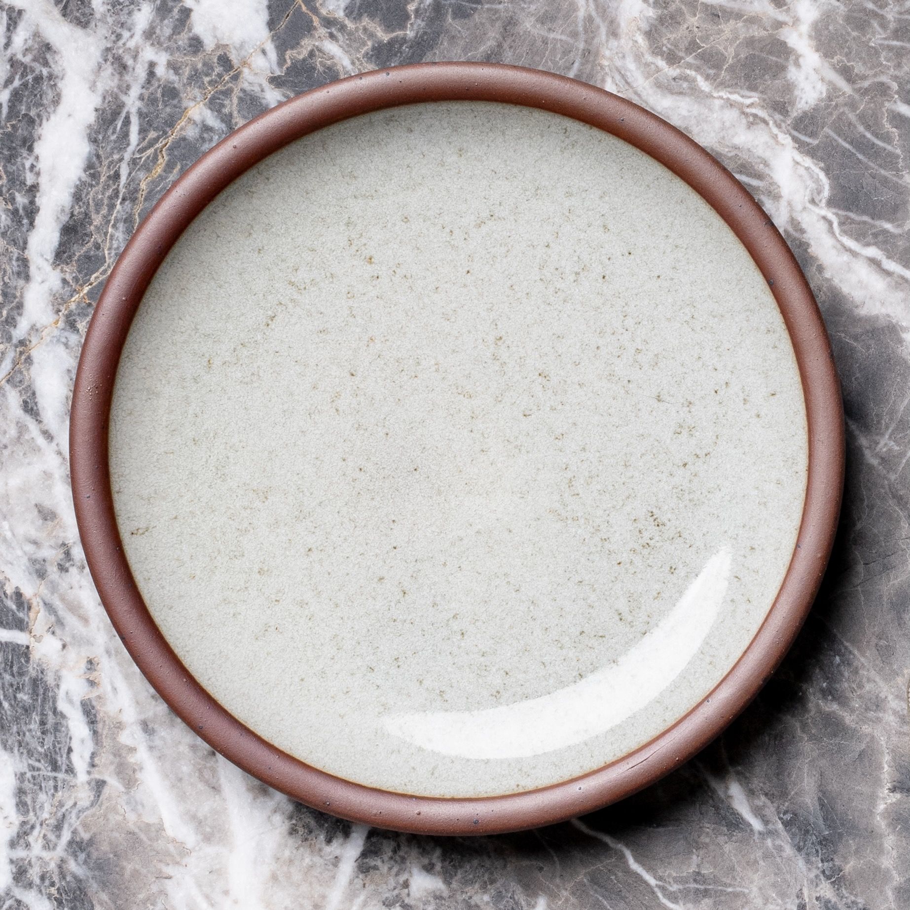 Round reflective icy white ceramic plate with an glazed rim on a gray marble surface.