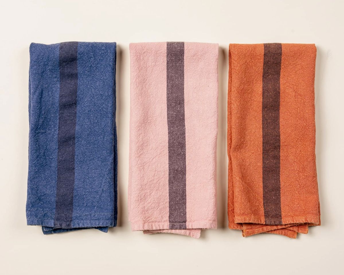 3 folded linen towels in an indigo, salmon, and terracotta color with a plum stripe in the middle of each