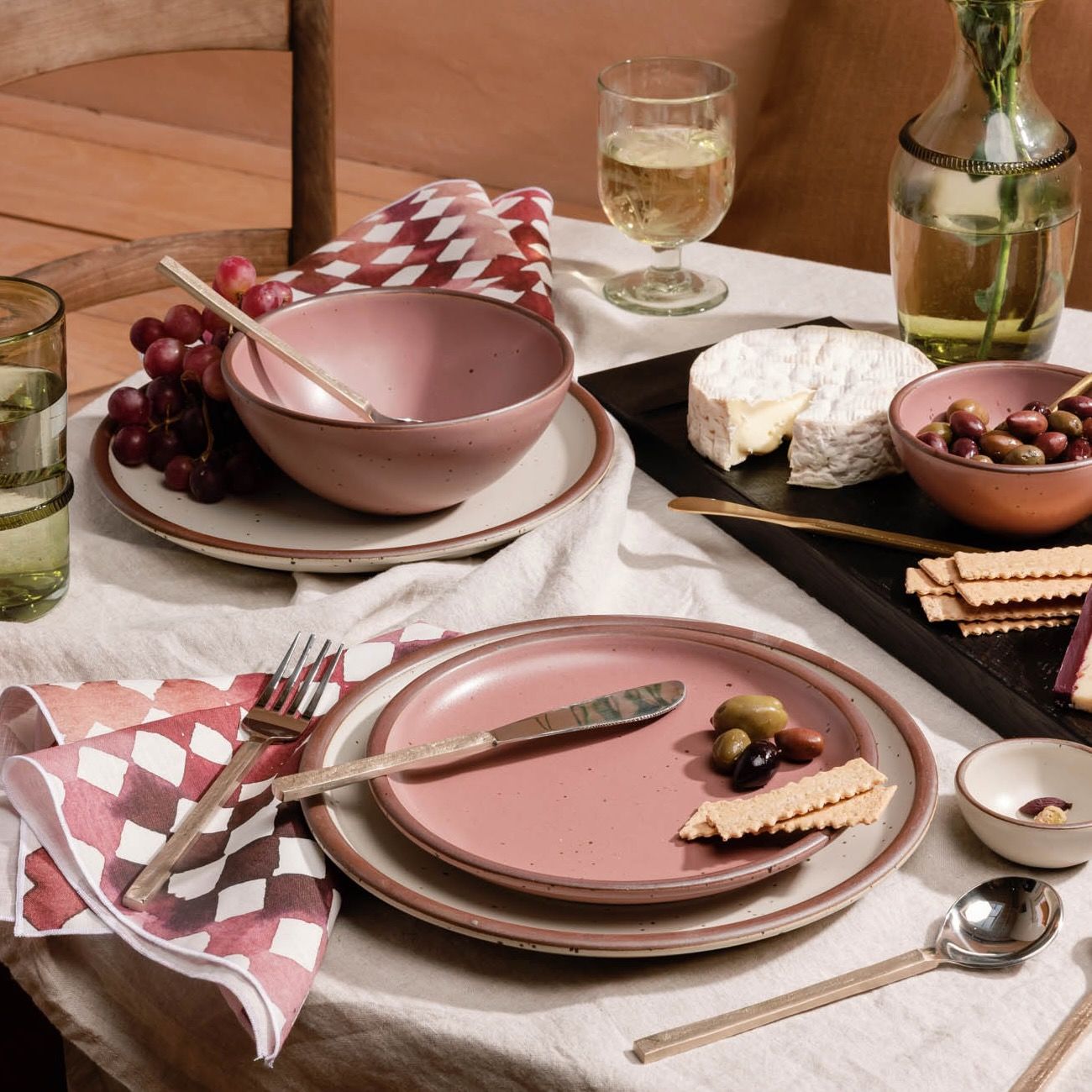 A table setting with ceramic dinnerware in cream and soft pink colors, patterned linens, a cheeseboard and more.