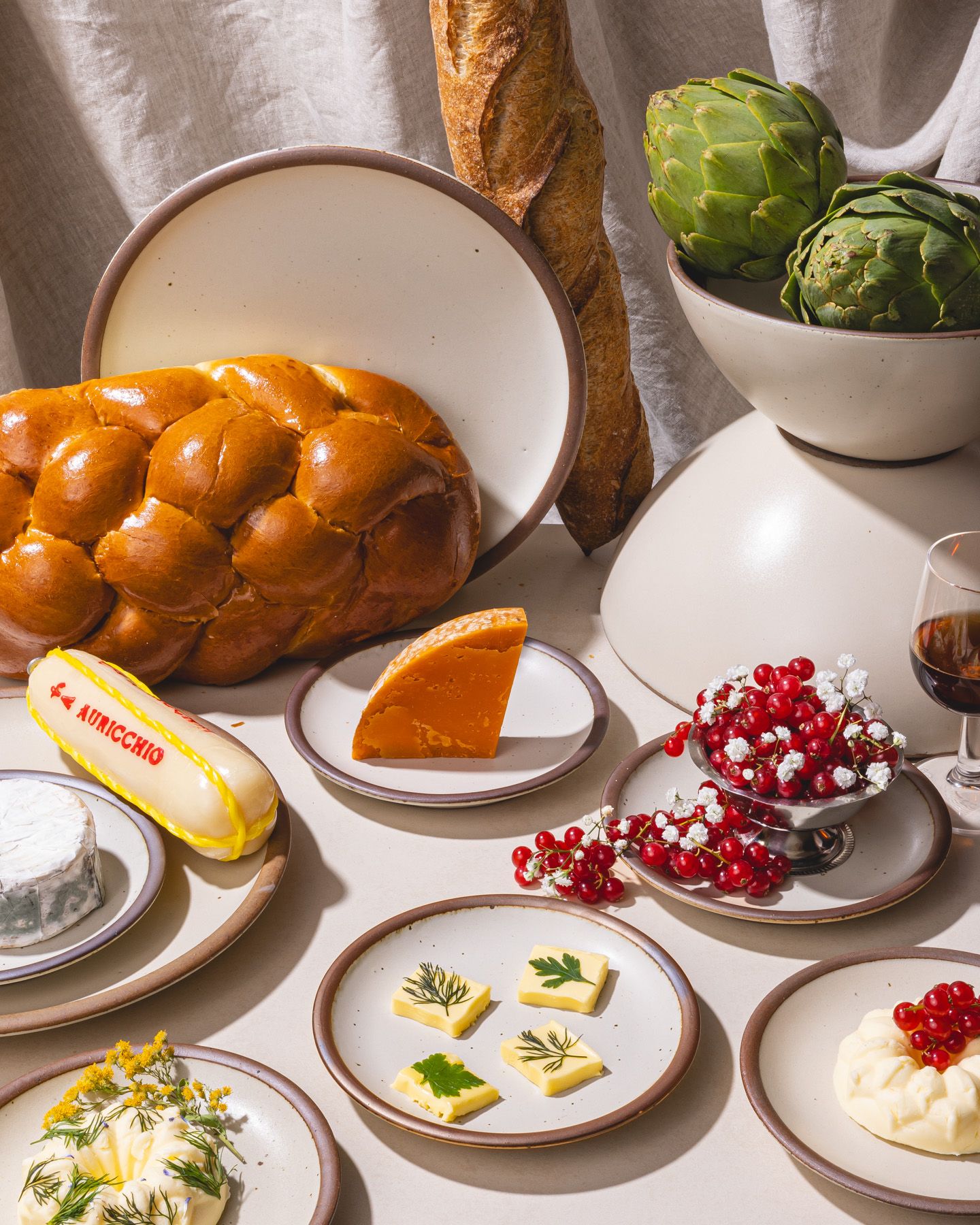 An artful display of cream ceramic plates with foods like berries, butter slices, bread, cheese, artichokes and more all around.