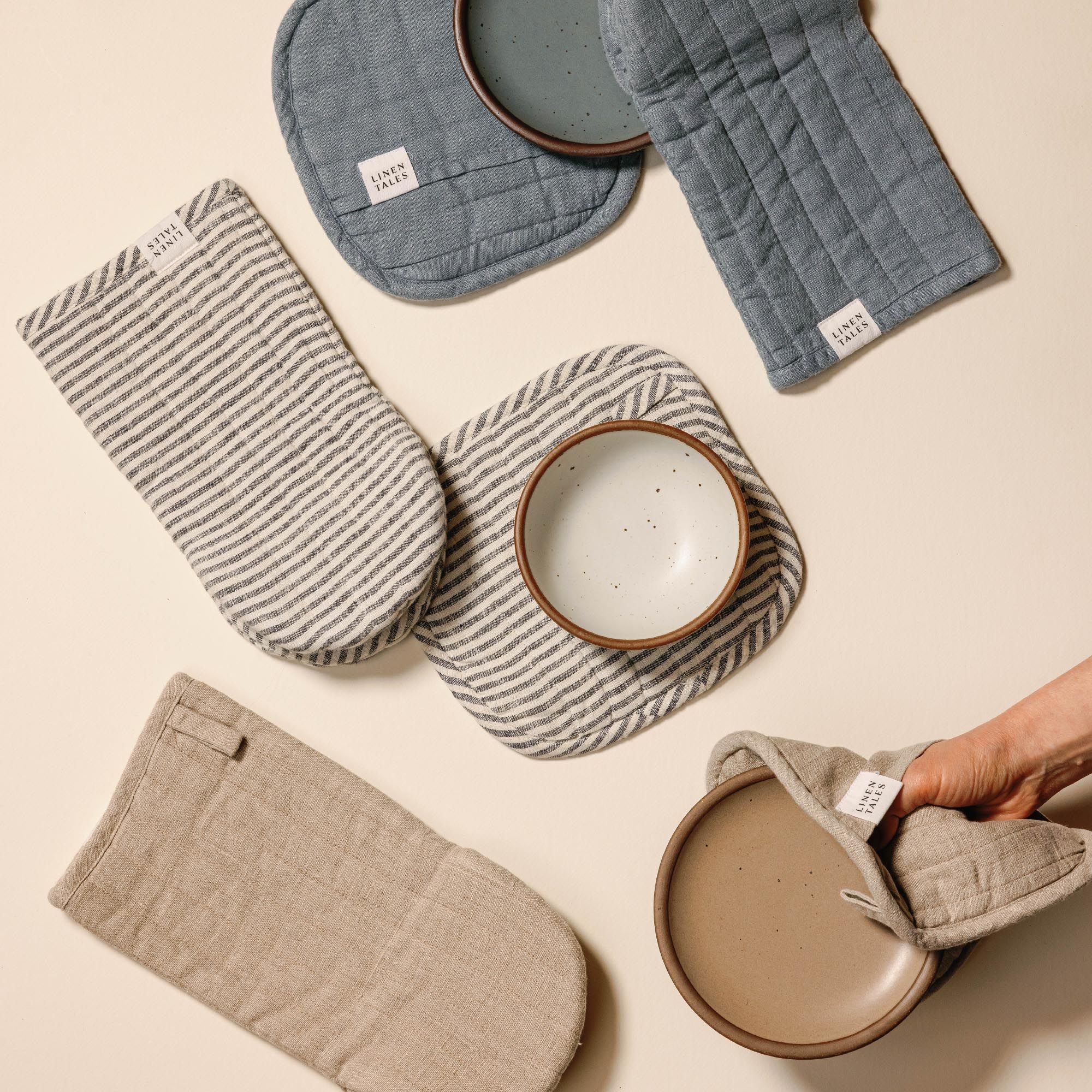 Quilted linen potholders and oven mitts in neutral beige, dusty blue, and navy-and-white stripes, each with a small ‘LINEN TALES’ label, arranged overlapping on a light background, with cake plates in matching colors.