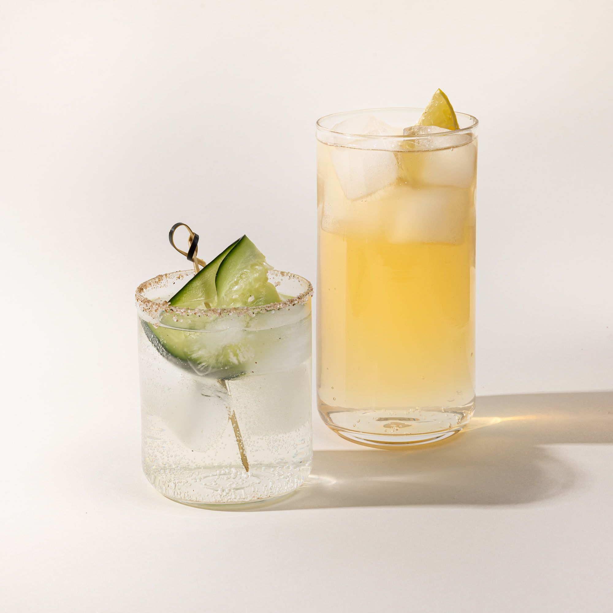 Two straight walled clear glasses - one tall, one short - filled with cocktails and fruit garnishes.