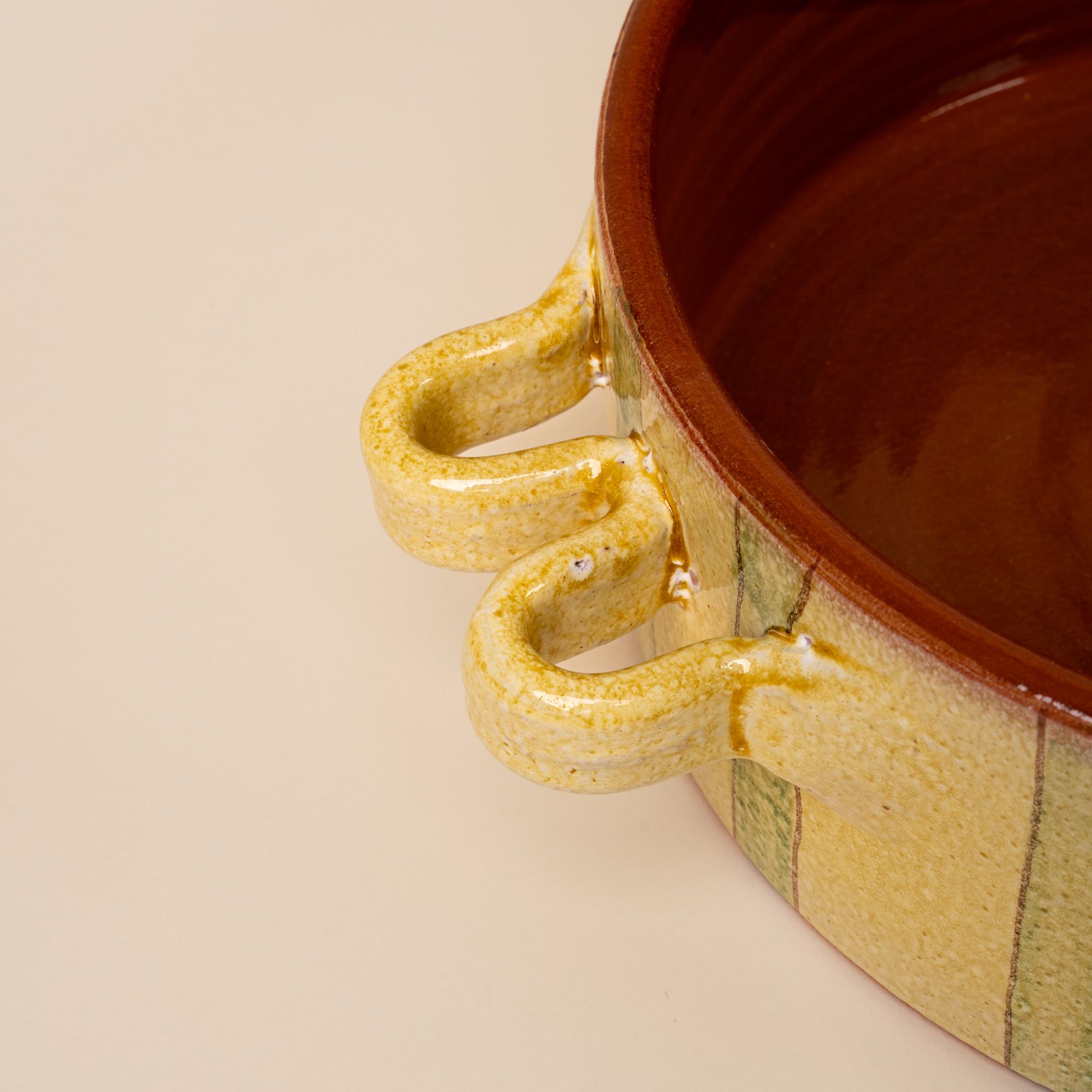 A closeup of the handle of a large terracotta casserole dish that is butter yellow and green striped with a terracotta interior.