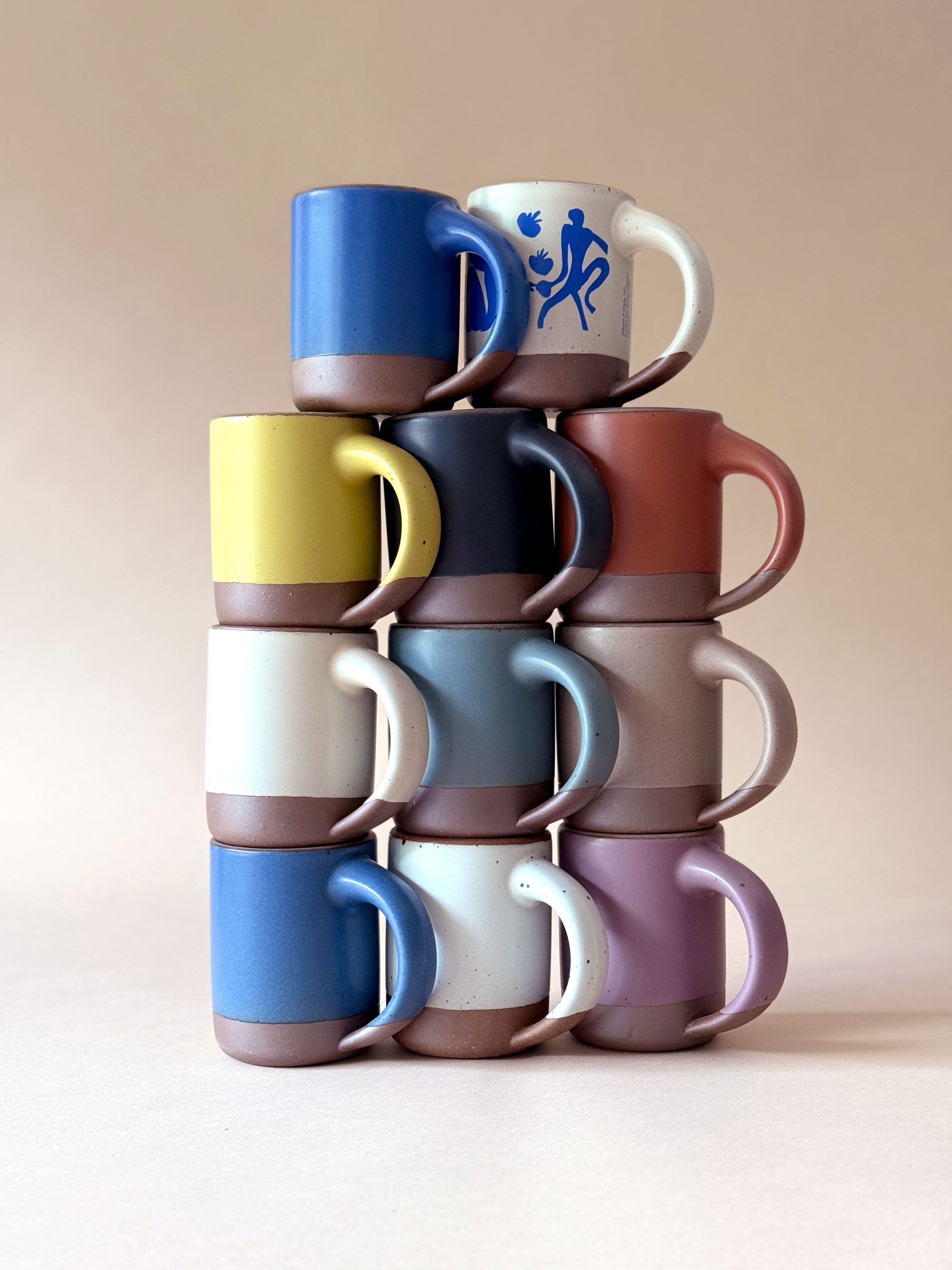 Stack of colorful ceramic mugs arranged in a pyramid, featuring a variety of solid glazes and one with blue abstract artwork.