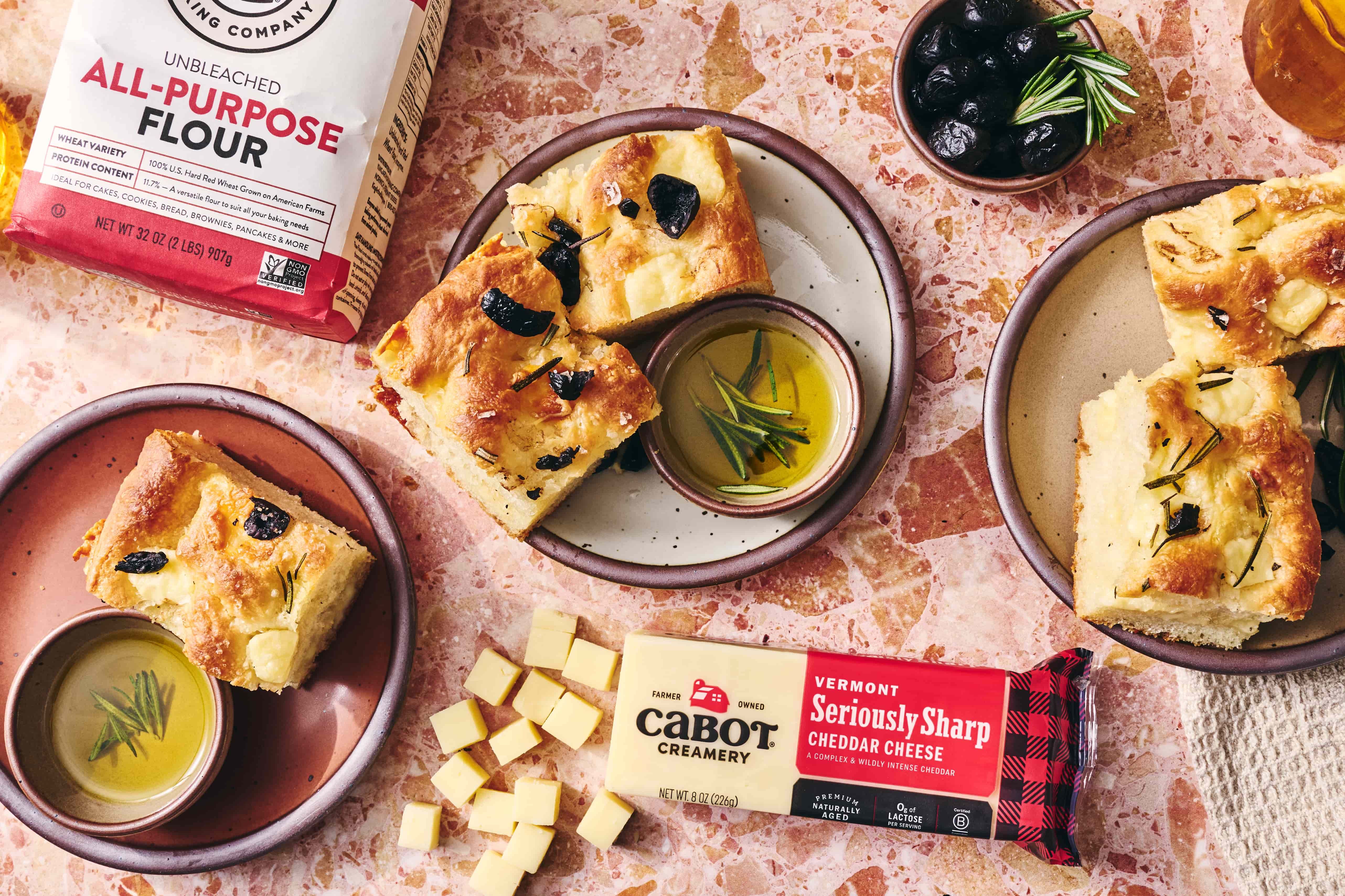 Slices of focaccia bread on ceramic cake plates with a bitty bowl filled with olive oil for dipping, artfully arranged next to Cabot Creamery cheese, and King Arthur Baking's flour.