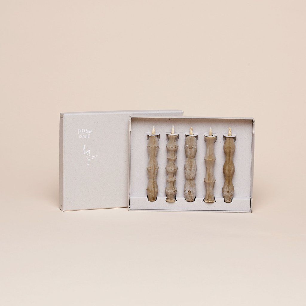 A rectangular box with lid behind it that contains five tan candles, taller than they are wide, each with a different pattern of flares and tapers
