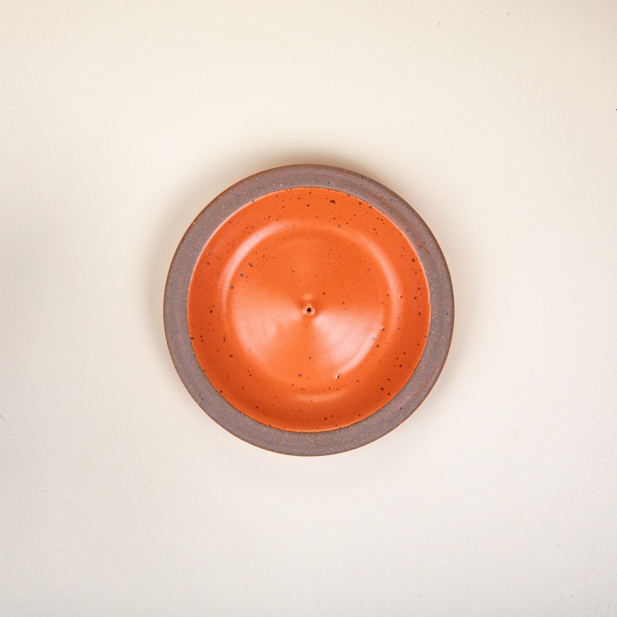 An overhead view of a ceramic plate-shaped incense stick holder with an unglazed rim and glazed in a bold orange color featuring lots of iron speckles.