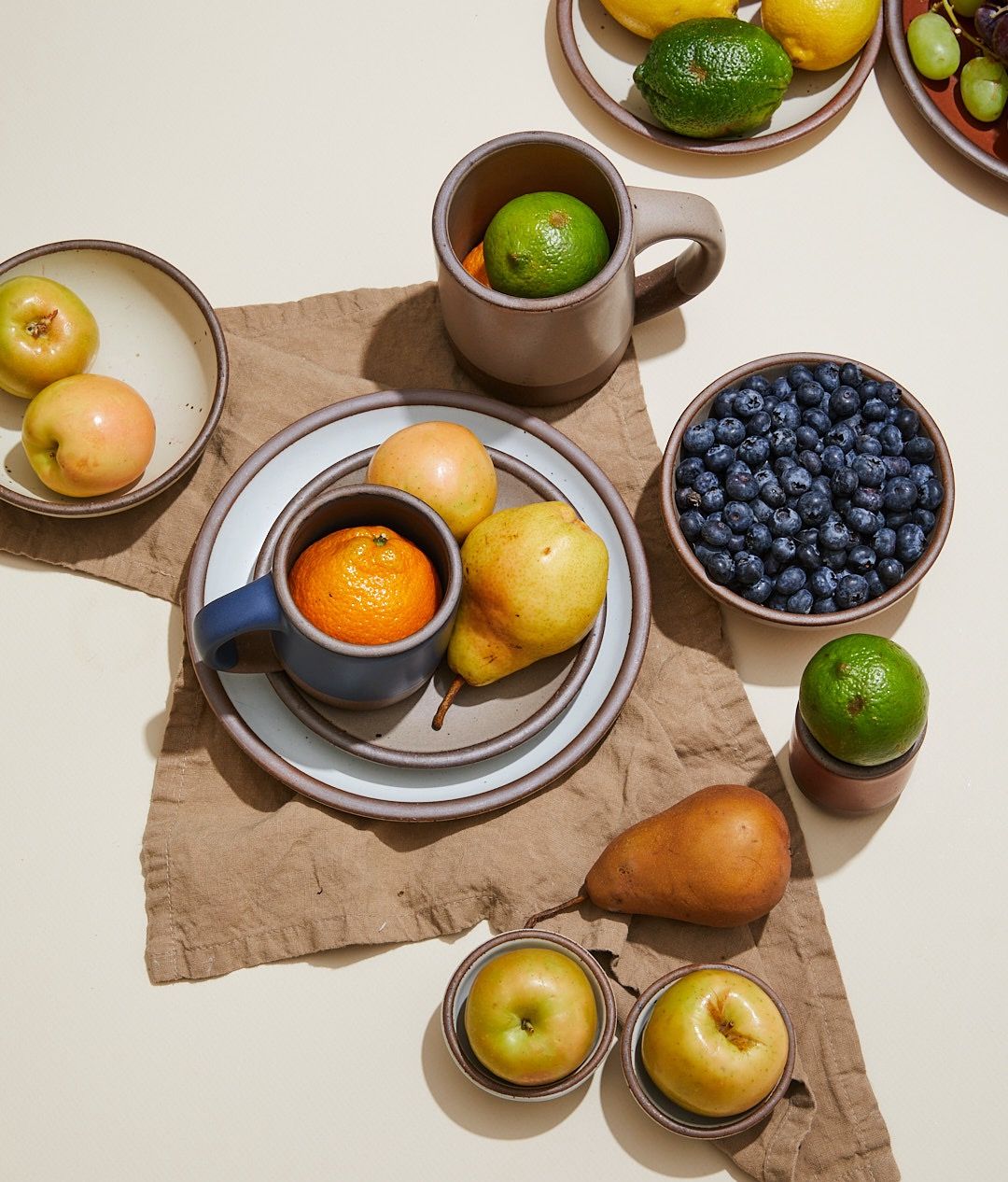 Overhead view of ceramic bowls, plates, and mugs in neutral colors filled with fruits and sitting on a linen napkin