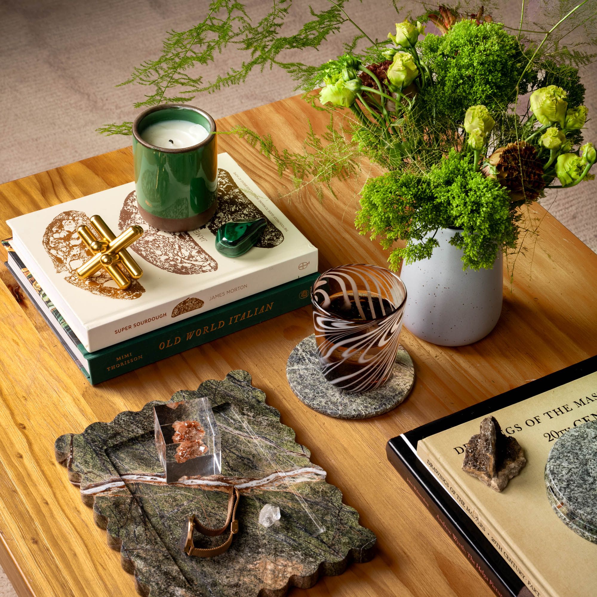 Coffee table styled with green candle, malachite stone, books, greenery in a white vase, scalloped marble tray, and patterned glass on coaster.