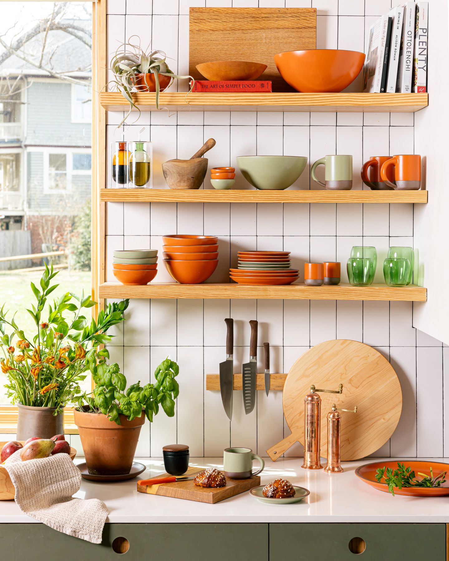 In a kitchen, ceramic bowls and mugs in bold orange and calming sage green colors fill floating shelves, along with green glasses and other kitchen tools. On the counter are various kitchen tools like a chef's knives, copper pepper and salt mills, cutting board with pastry, and potted plants.
