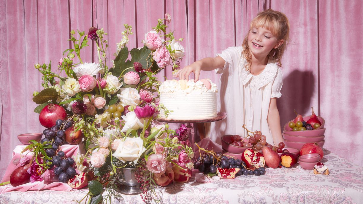 A child reaches for a decadent cake sitting on a colorful table with lots of flowers, fruits, and dishes in a pink color, all with a pink background
