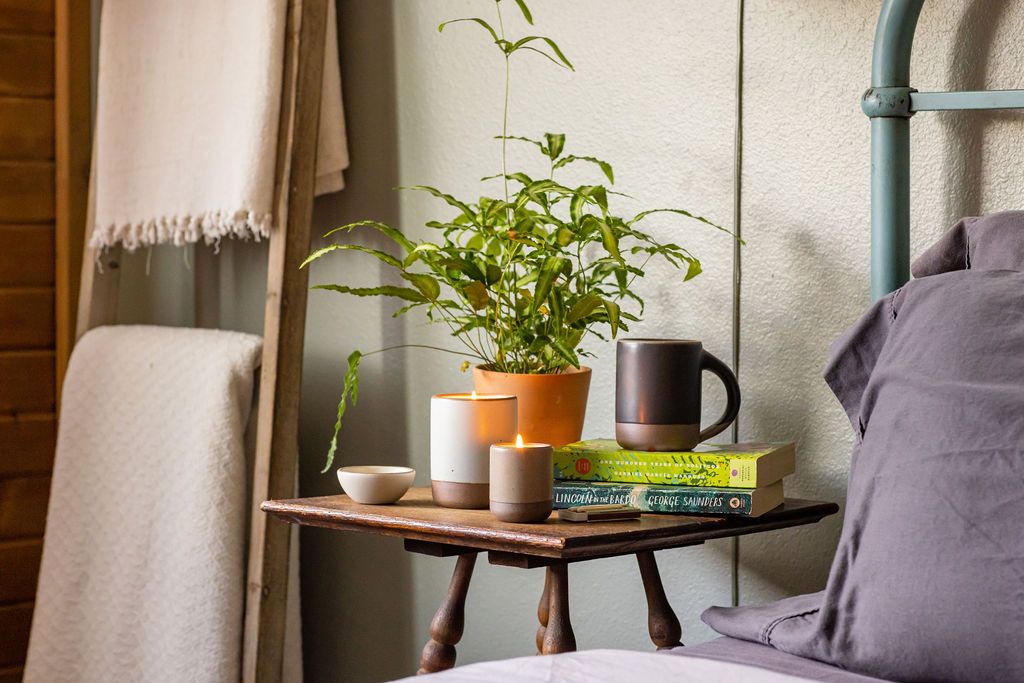 On a bedside table, sits 2 lit candles, an East Fork Mug in Black Mountain, a small stack of books, and a potted plant.