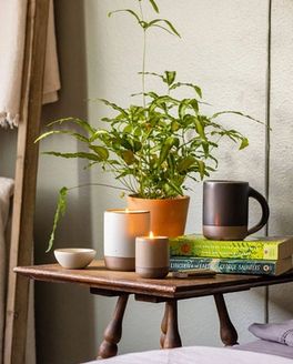 On a bedside table, sits 2 lit candles, an East Fork Mug in Black Mountain, a small stack of books, and a potted plant.