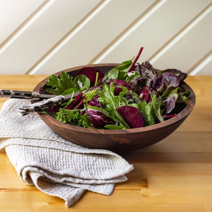 A large wooden bowl filled with salad and a serving fork and spoon on top. To the right of the bowl is a natural kitchen towel.