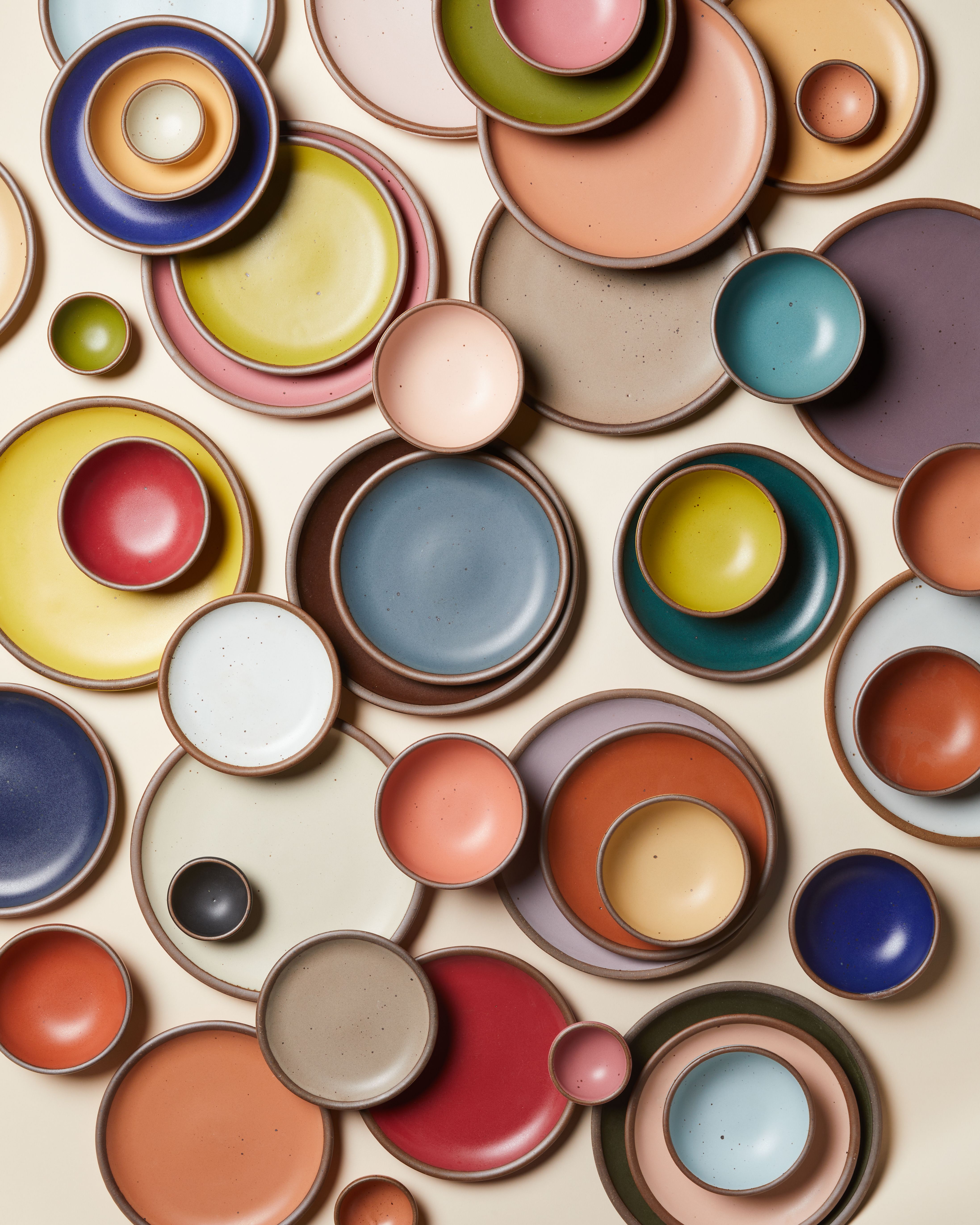 A person sporting a high bun in a shop jumpsuit stands in front of a colorful wall of hanging plates.