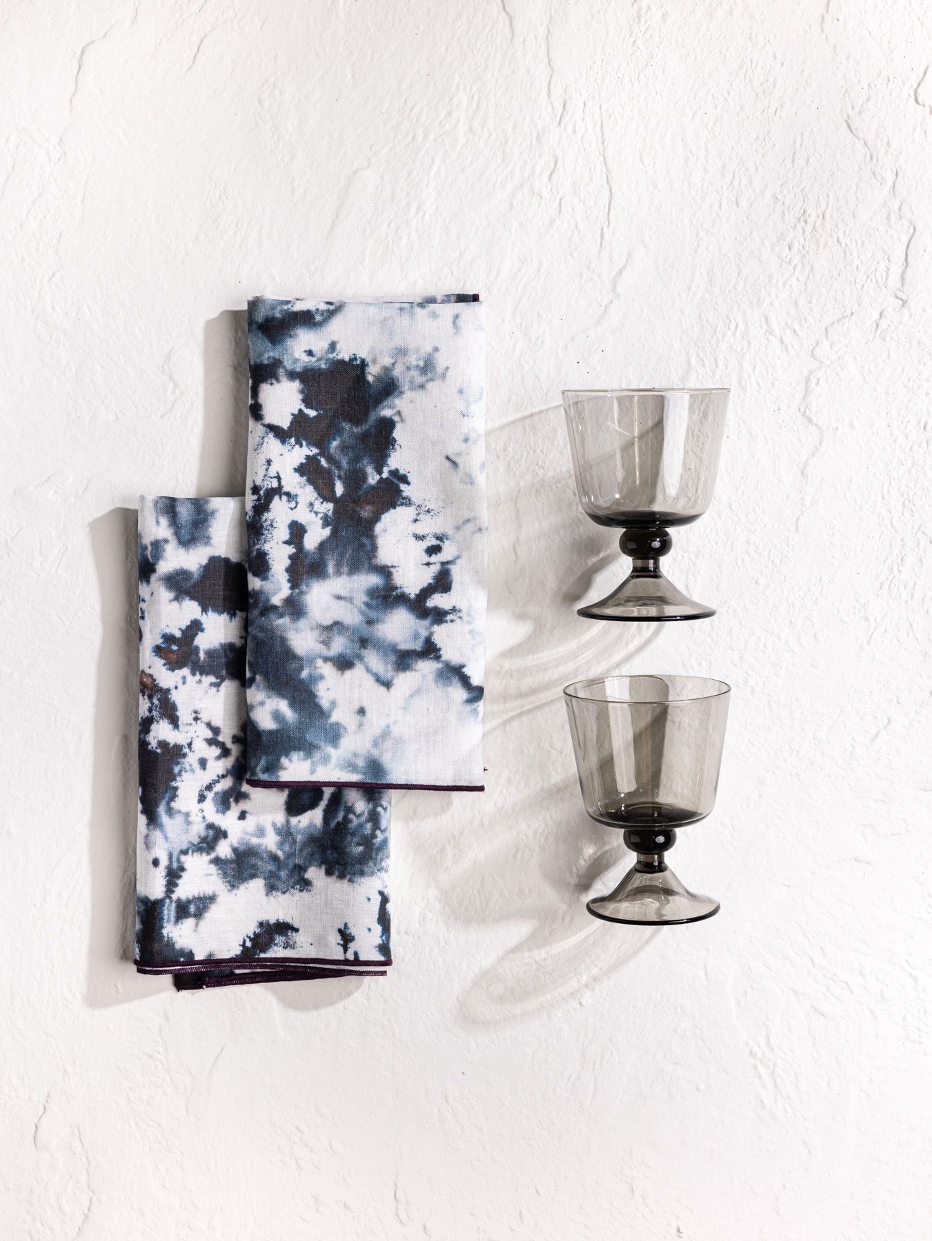 Top-down view of two dark indigo and white marbled napkins folded into rectangles and two smoky gray glasses with stout stems.